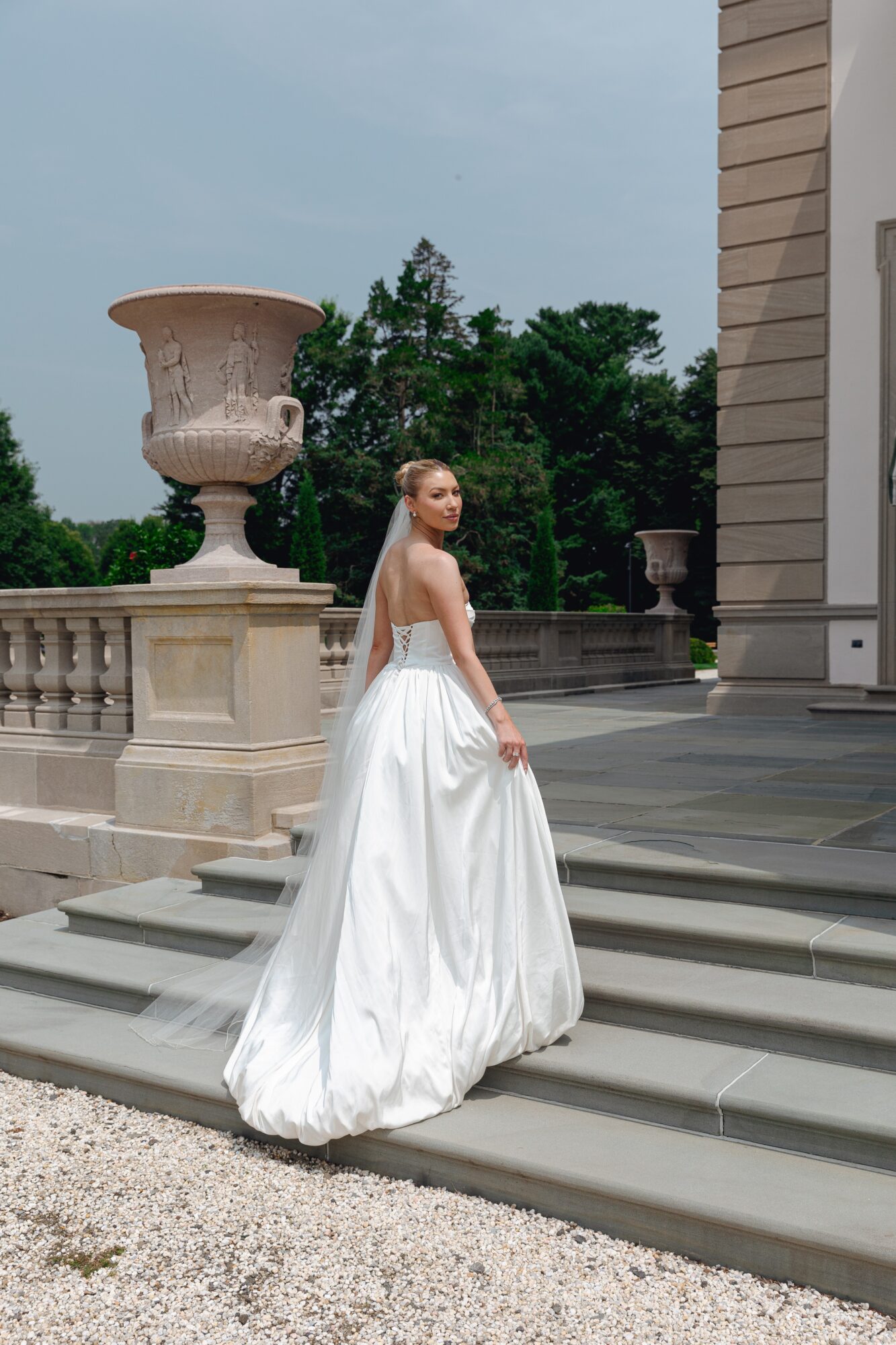 Woman in a white gown standing on steps outside a building with columns and large urns, trees in background.
