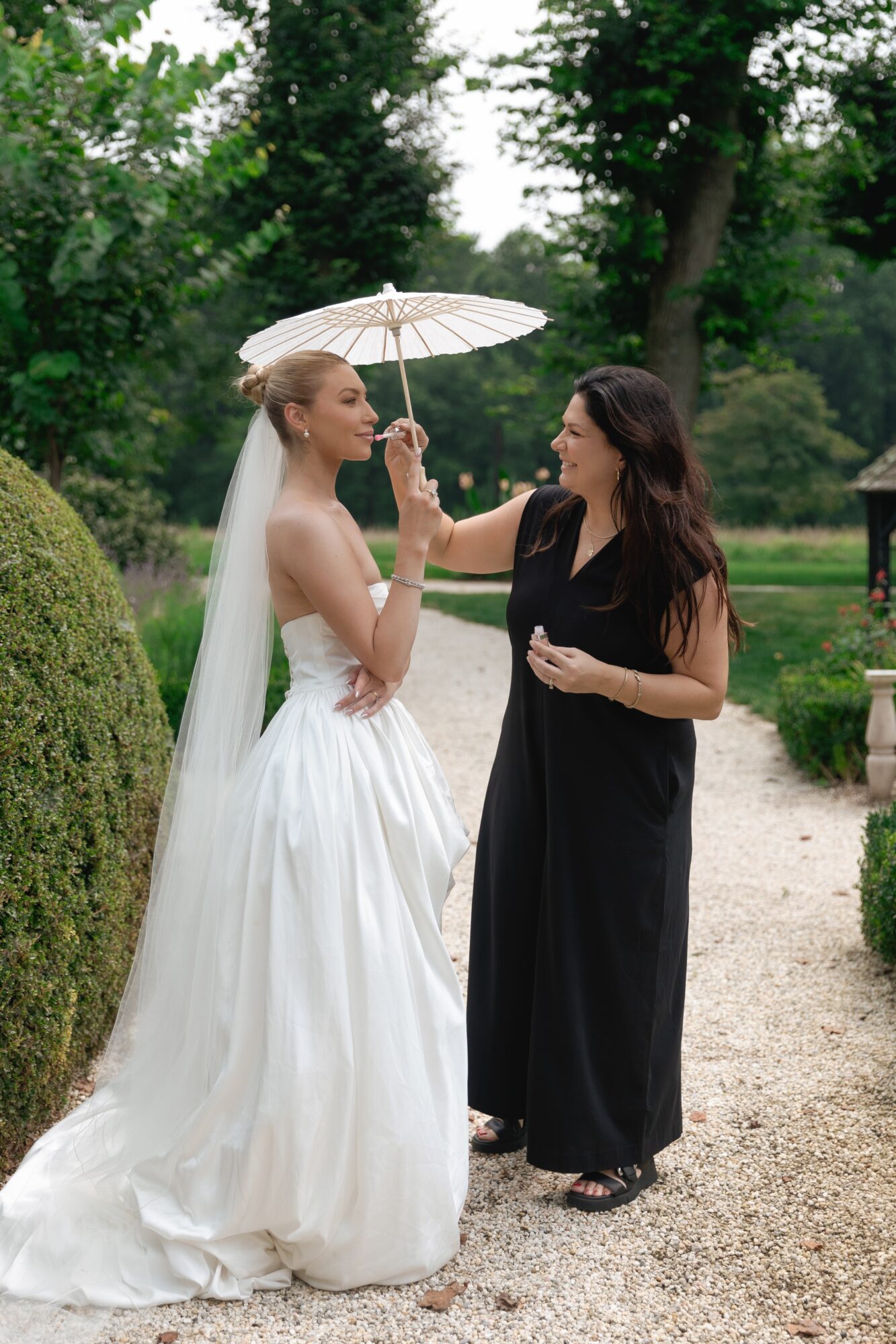 Bride in wedding dress and veil holding umbrella, talking to woman in black dress outdoors, greenery in background.