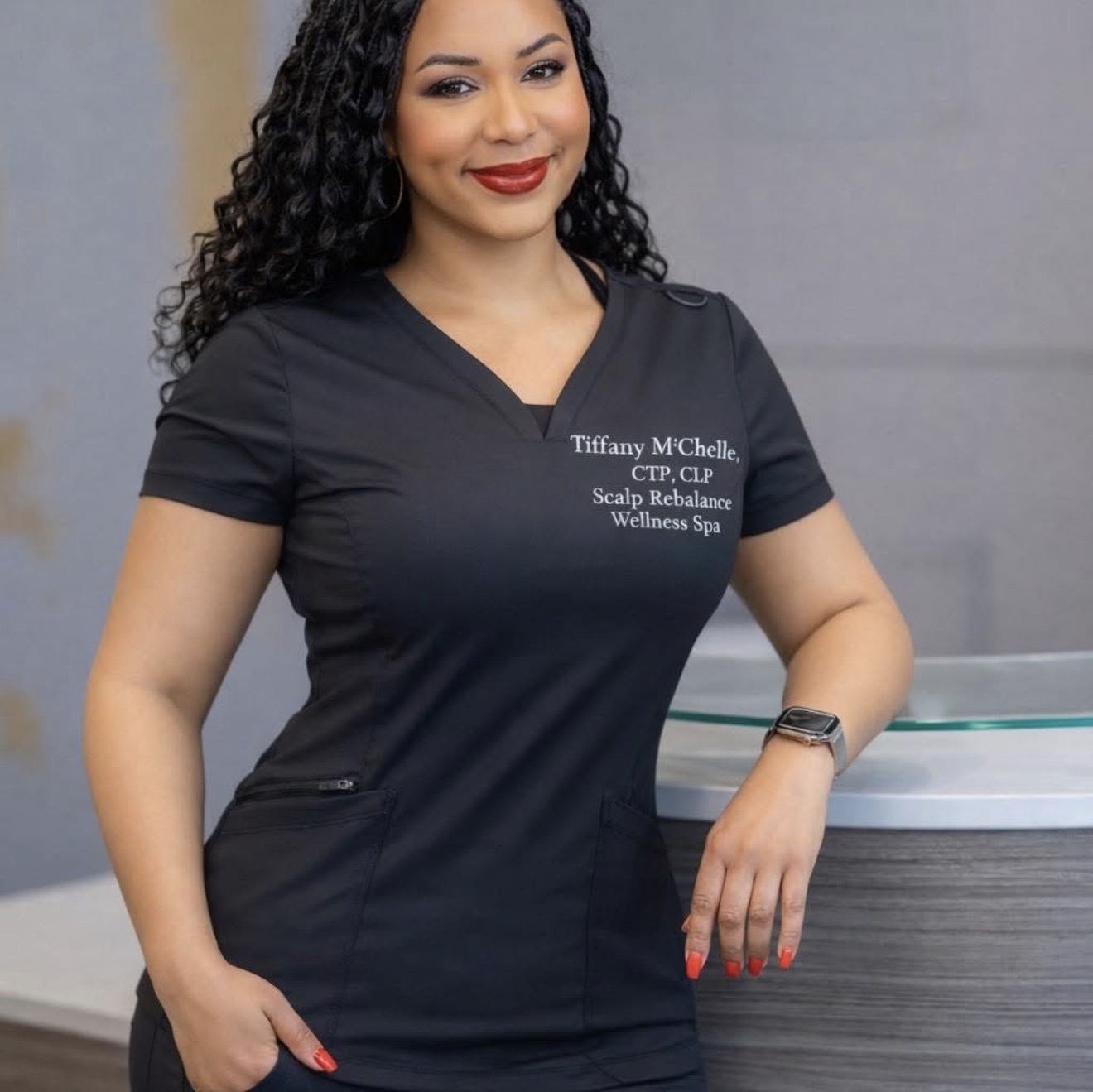 Woman with curly hair in black uniform standing at reception desk, smiling, with text on uniform, wellness spa setting.