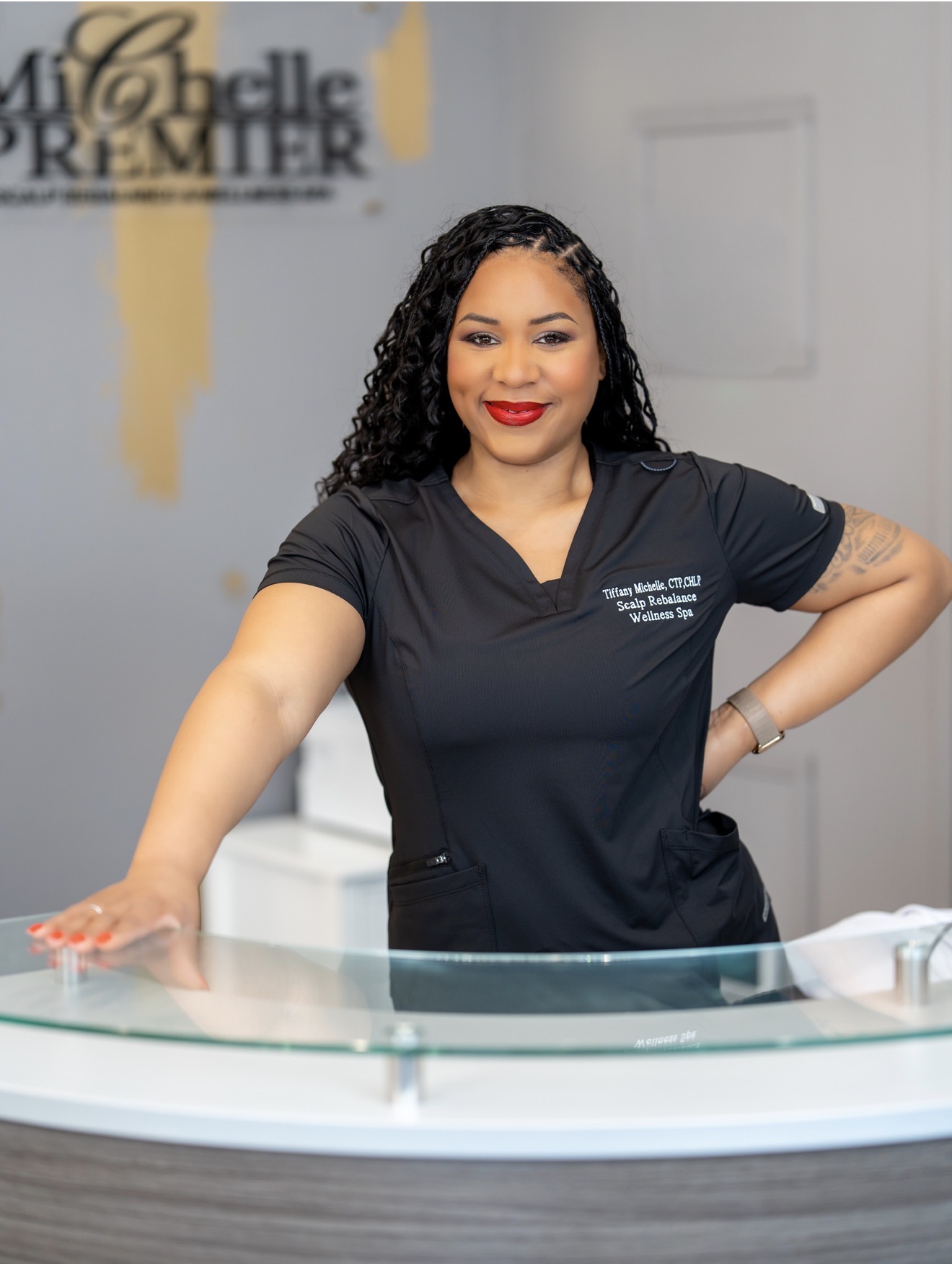 Woman with curly hair and red lipstick standing behind a glass counter, smiling, in a professional uniform inside a clinic or office.
