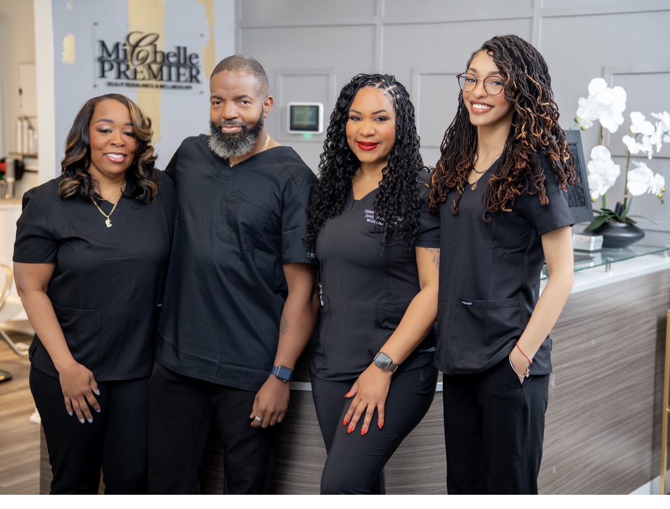 Four people in black scrubs standing together in a medical office, smiling, with a reception desk and orchids in the background.