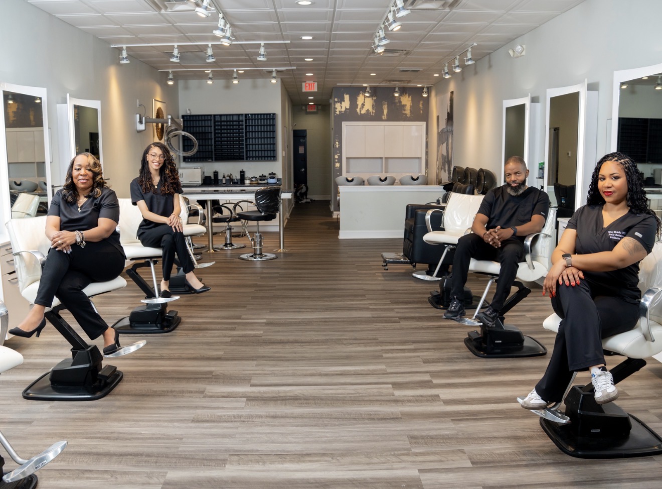 Five people sit on chairs in a spacious, well-lit room with wooden flooring and white walls, spaced apart.