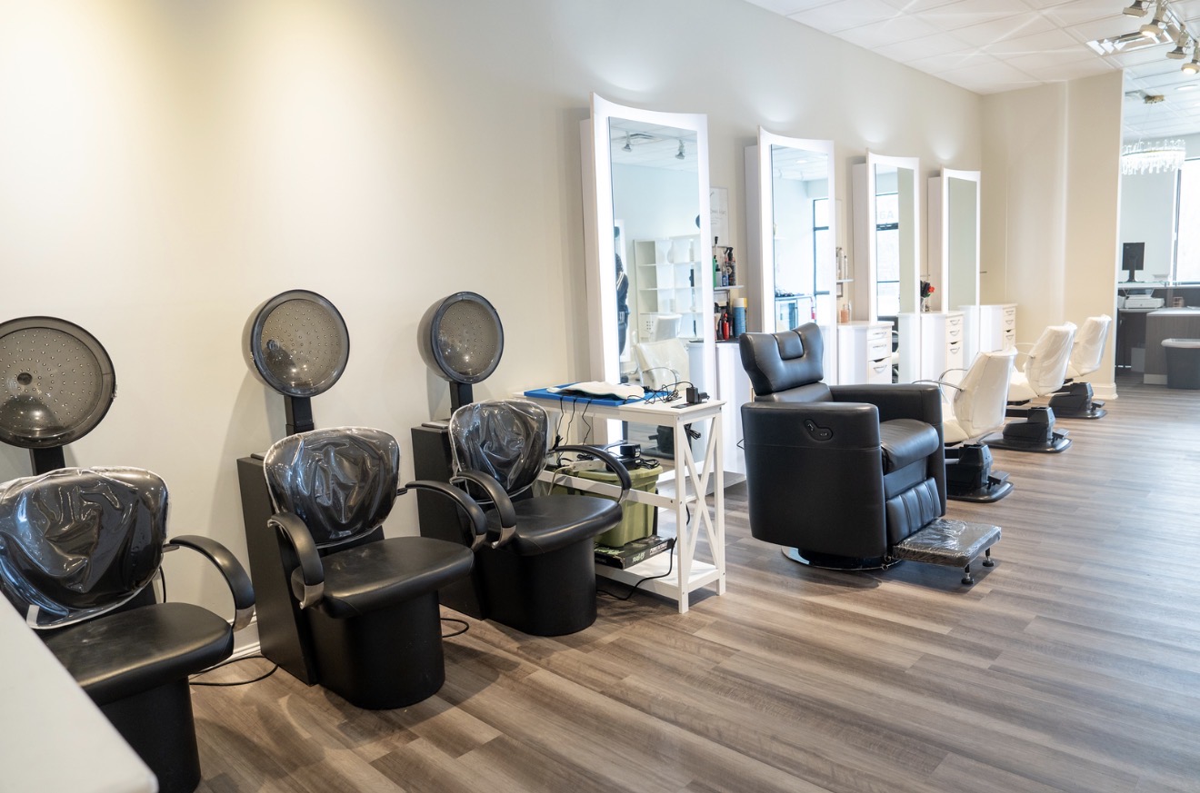 Salon with black chairs, hairdryers, and mirrors, wooden floor, and white walls, with additional seating in the background.