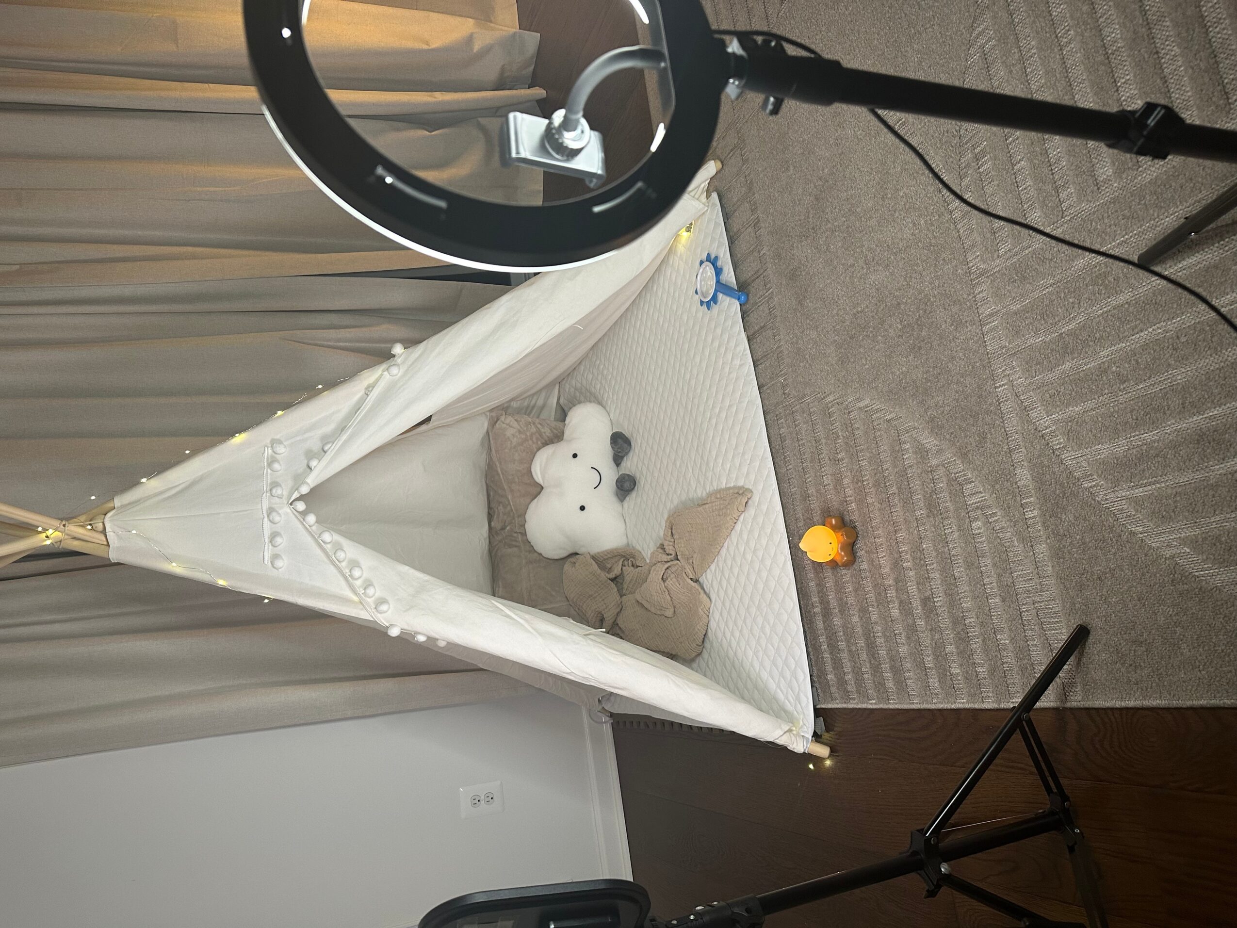 A small bed with a teddy bear and a plush toy inside, set on a carpeted floor with a ring light and tripod nearby.