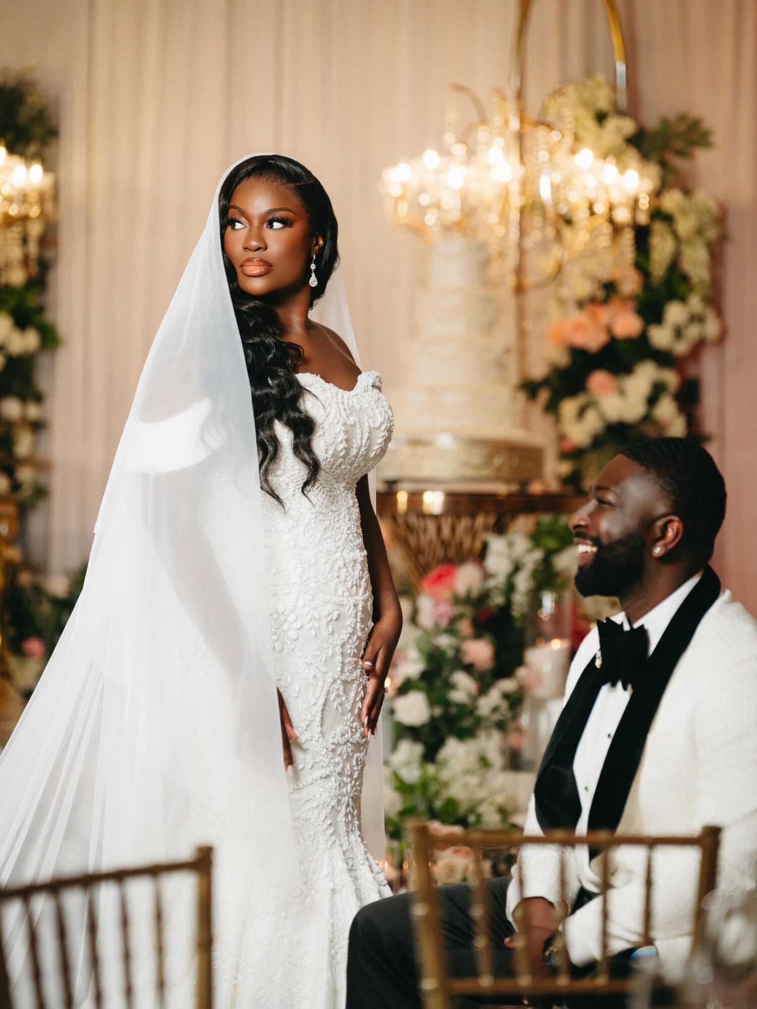 Bride in a white wedding dress and veil stands with a groom in a tuxedo, in a decorated venue with flowers and chandeliers.