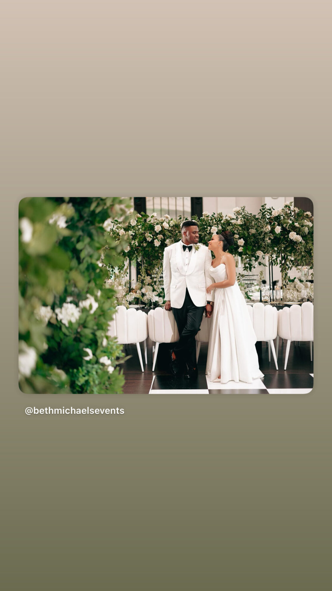 A couple dressed in wedding attire standing together at a decorated event with greenery and white flowers.