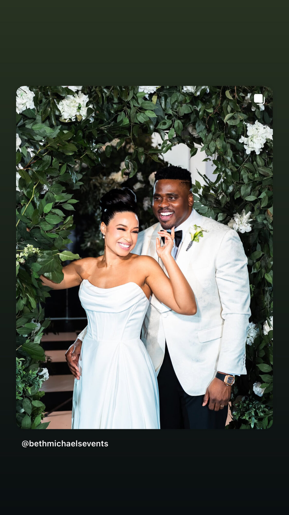 A smiling couple in formal attire standing outdoors with white flowers and greenery behind them.