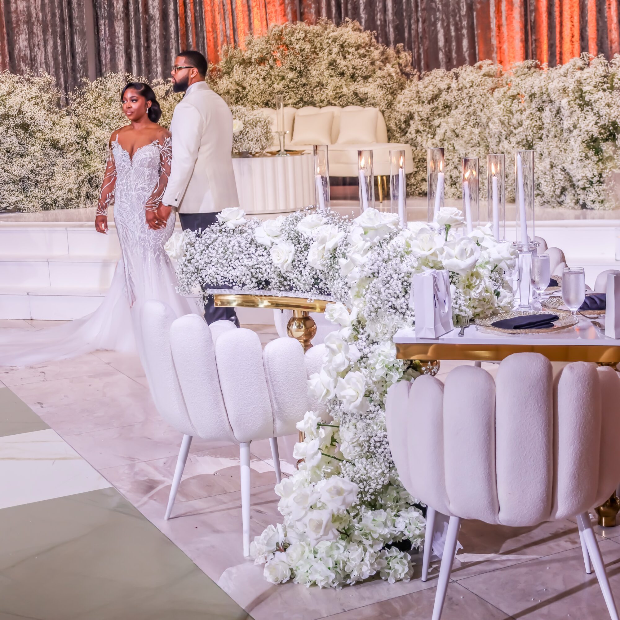 A couple stands holding hands at a decorated wedding reception with floral arrangements and elegant table settings.