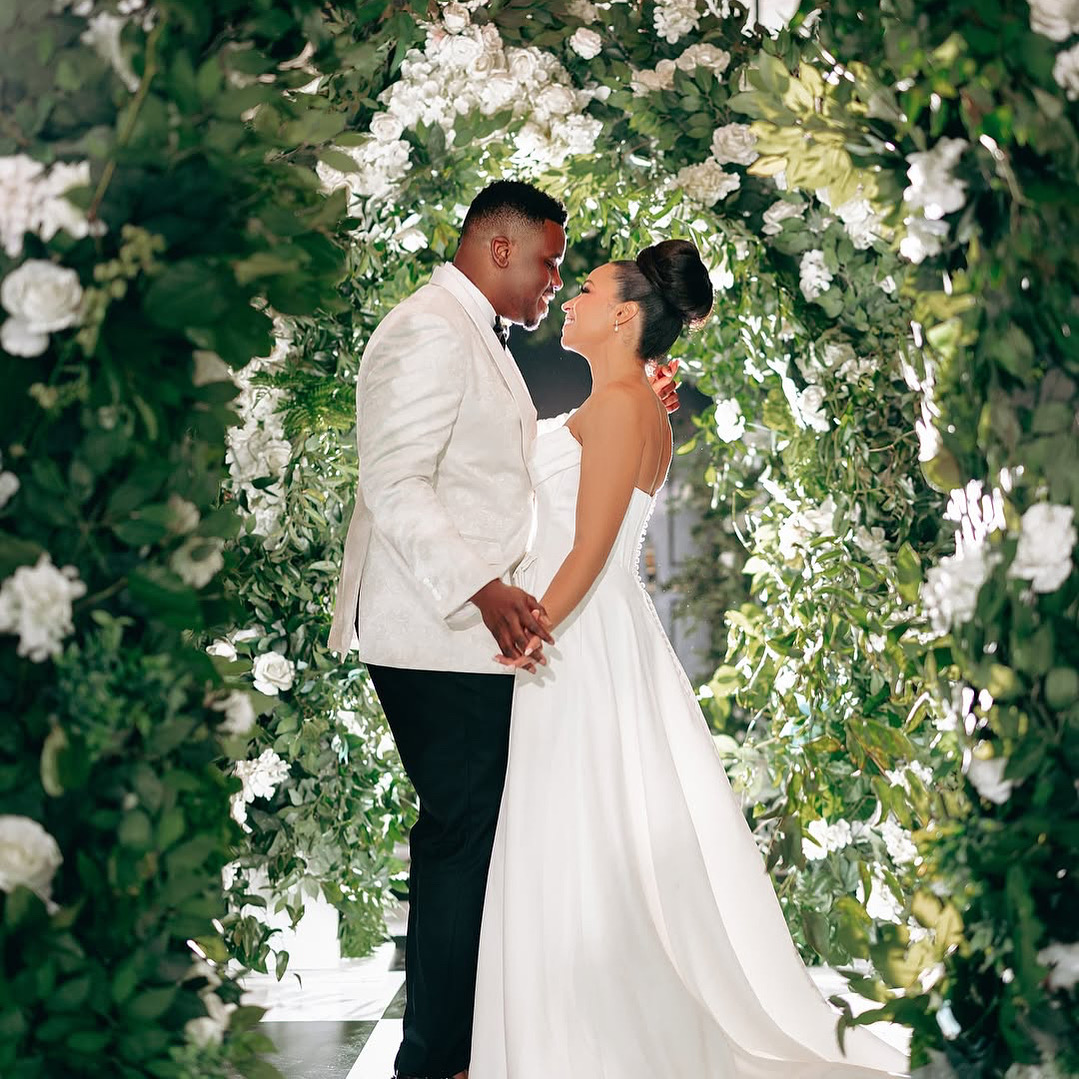 Couple in wedding attire holding hands and facing each other in front of a floral archway, smiling.