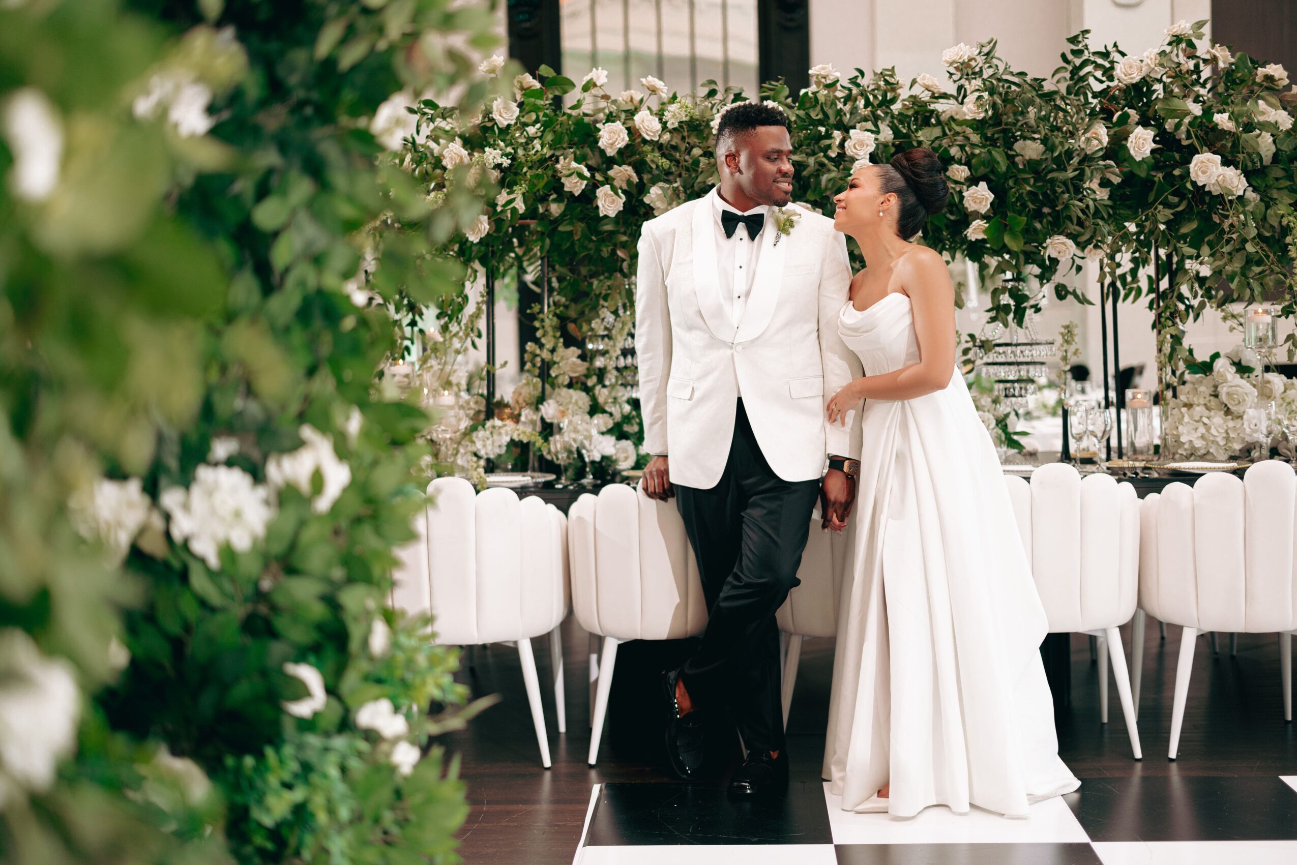 A bride and groom stand together at a wedding reception, holding hands, with floral decorations and a table in the background.