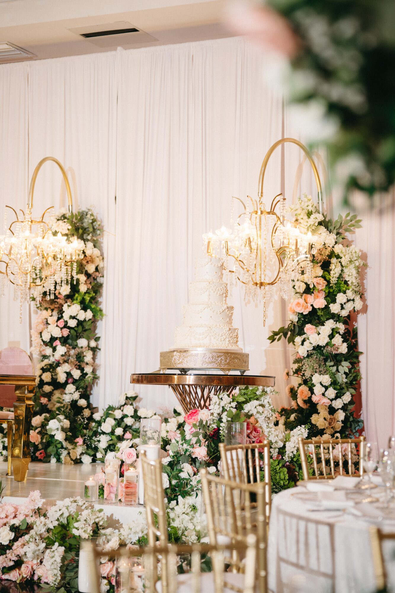Decorative floral arrangements with gold stands and a cake on a table in a decorated event space.