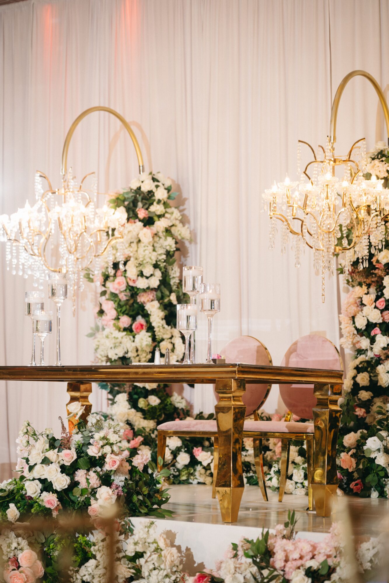 Decorative floral arrangements with white and pink flowers, gold accents, and chairs on a stage with a white curtain backdrop.