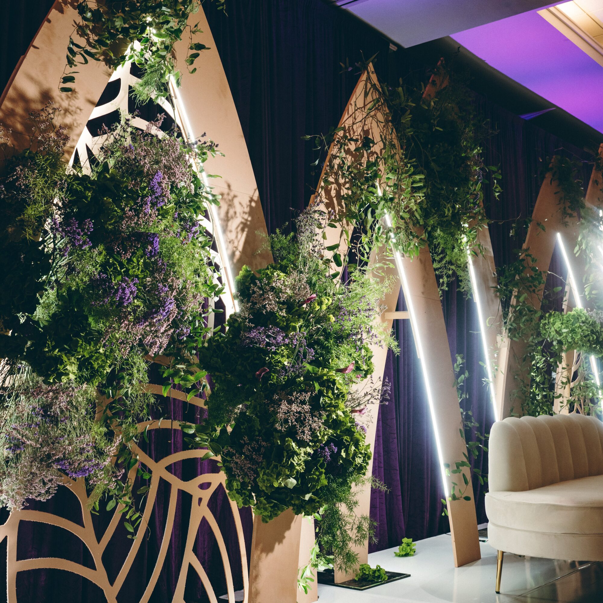 Decorative display with lush green and purple flowers, plants, and draped dark curtains, with a beige chair in the foreground.