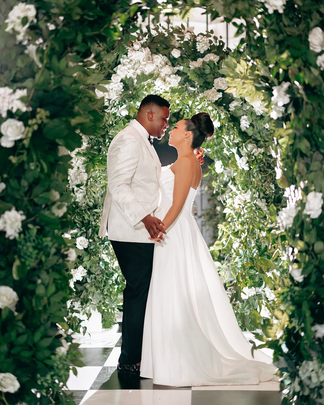 A bride and groom stand close, holding hands, in front of a floral arch on a checkered floor, smiling at each other.