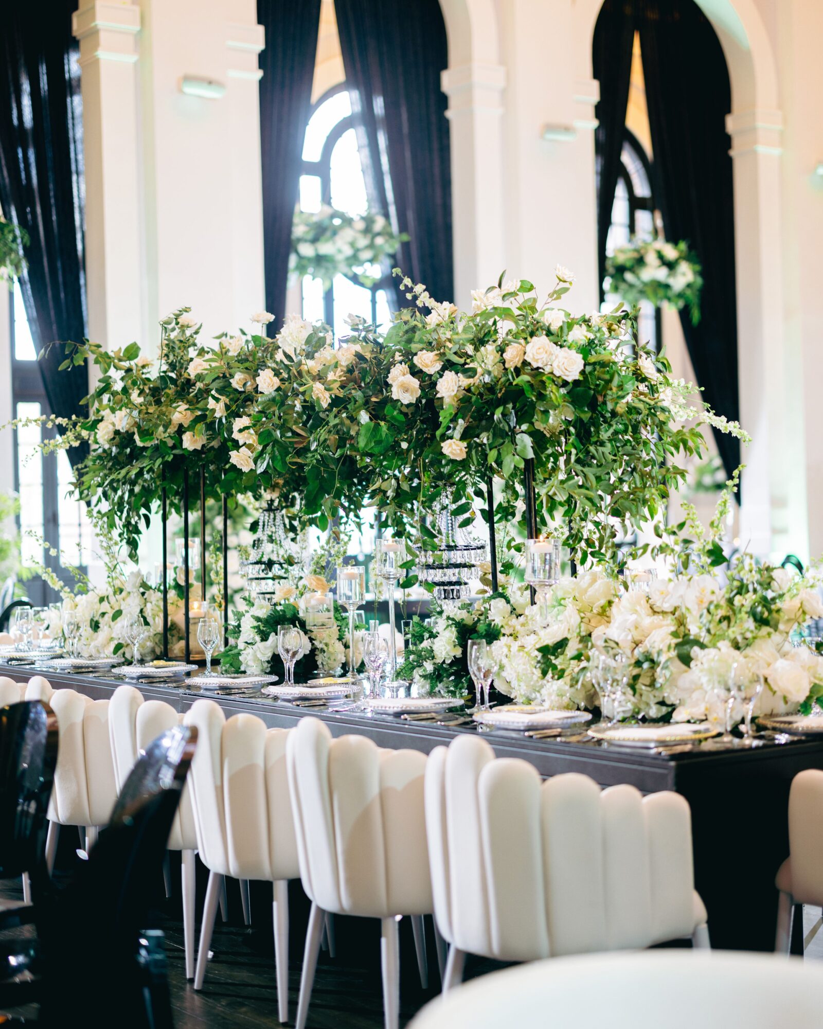 Elegant table setup with white floral arrangements and white chairs in a bright, ornate room with large windows.