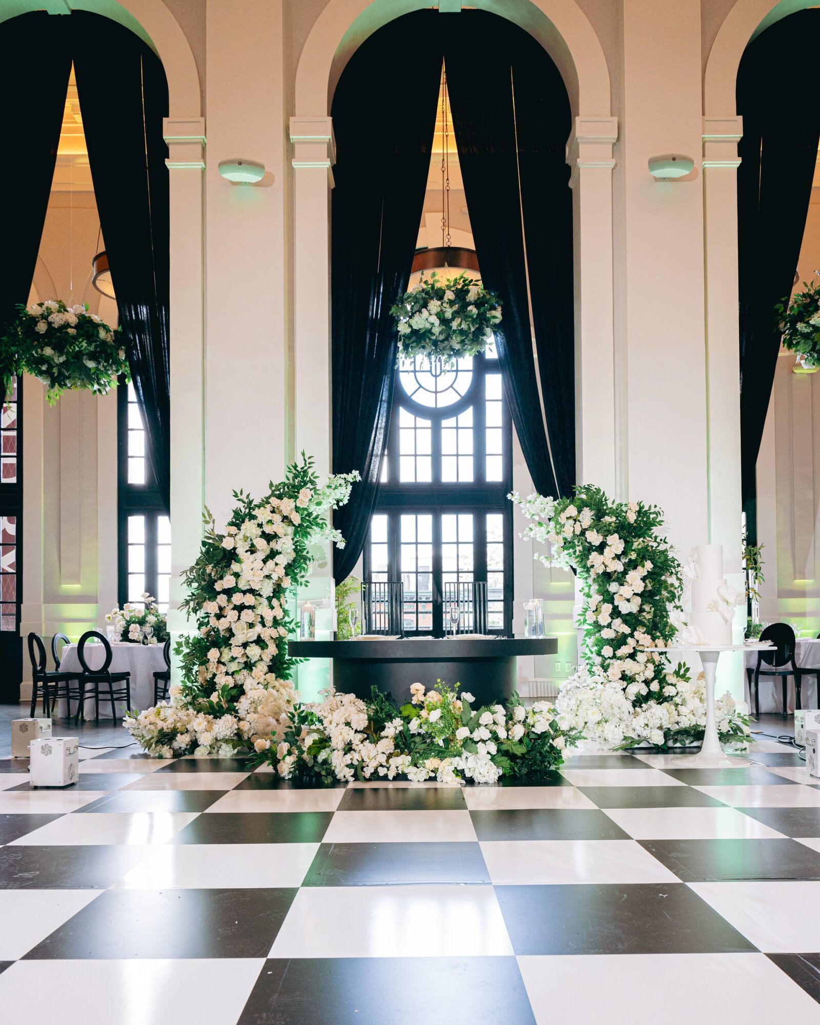 Elegant indoor space with black and white checkered floor, floral arrangements, and large windows with dark curtains.
