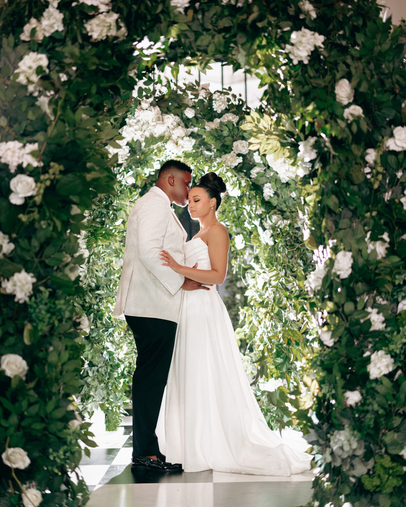 Couple in wedding attire standing close under a floral arch, surrounded by greenery and white flowers, in a garden setting.