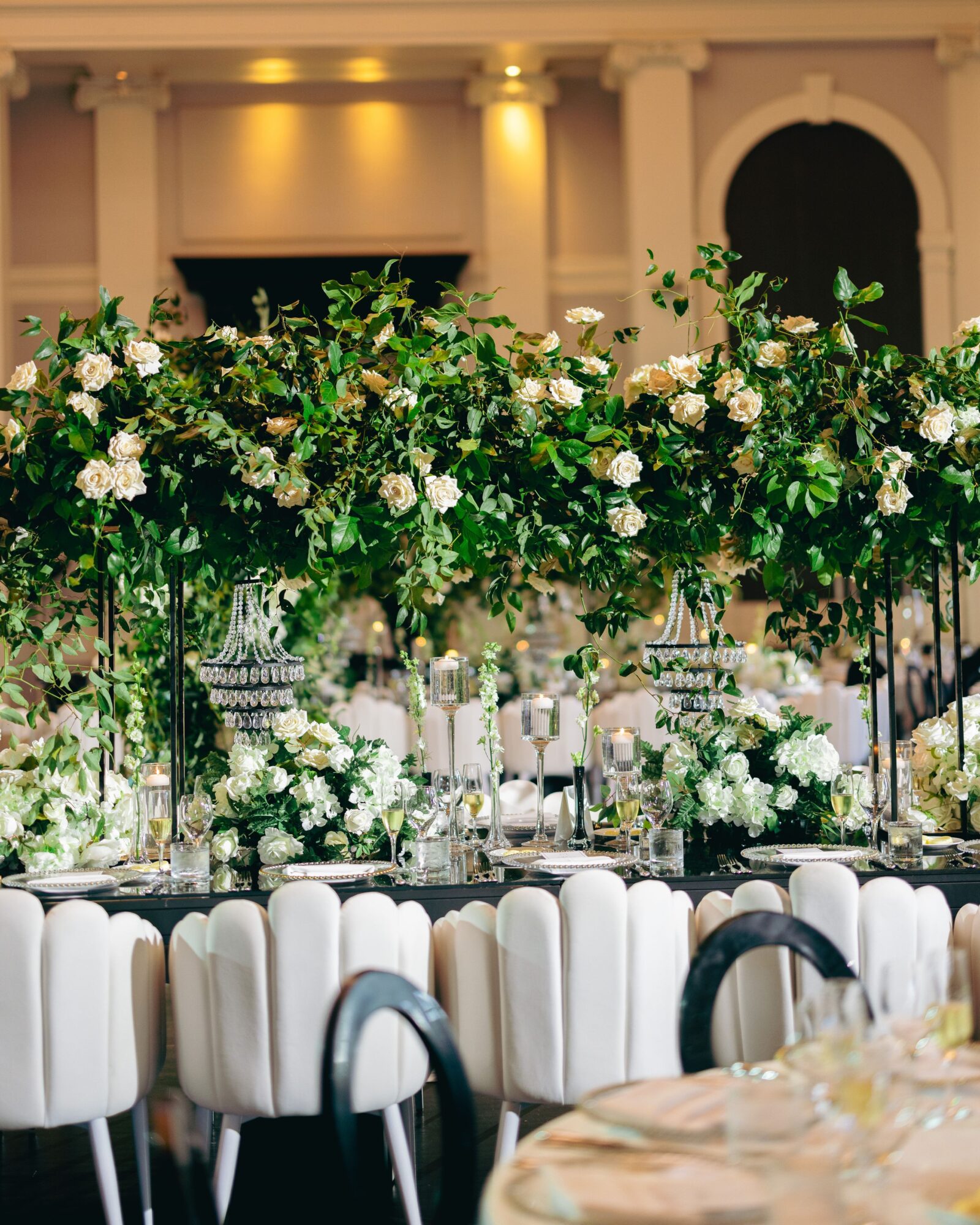 Elegant banquet table with white chairs, floral centerpiece with white and green flowers, set in a grand hall with columns.