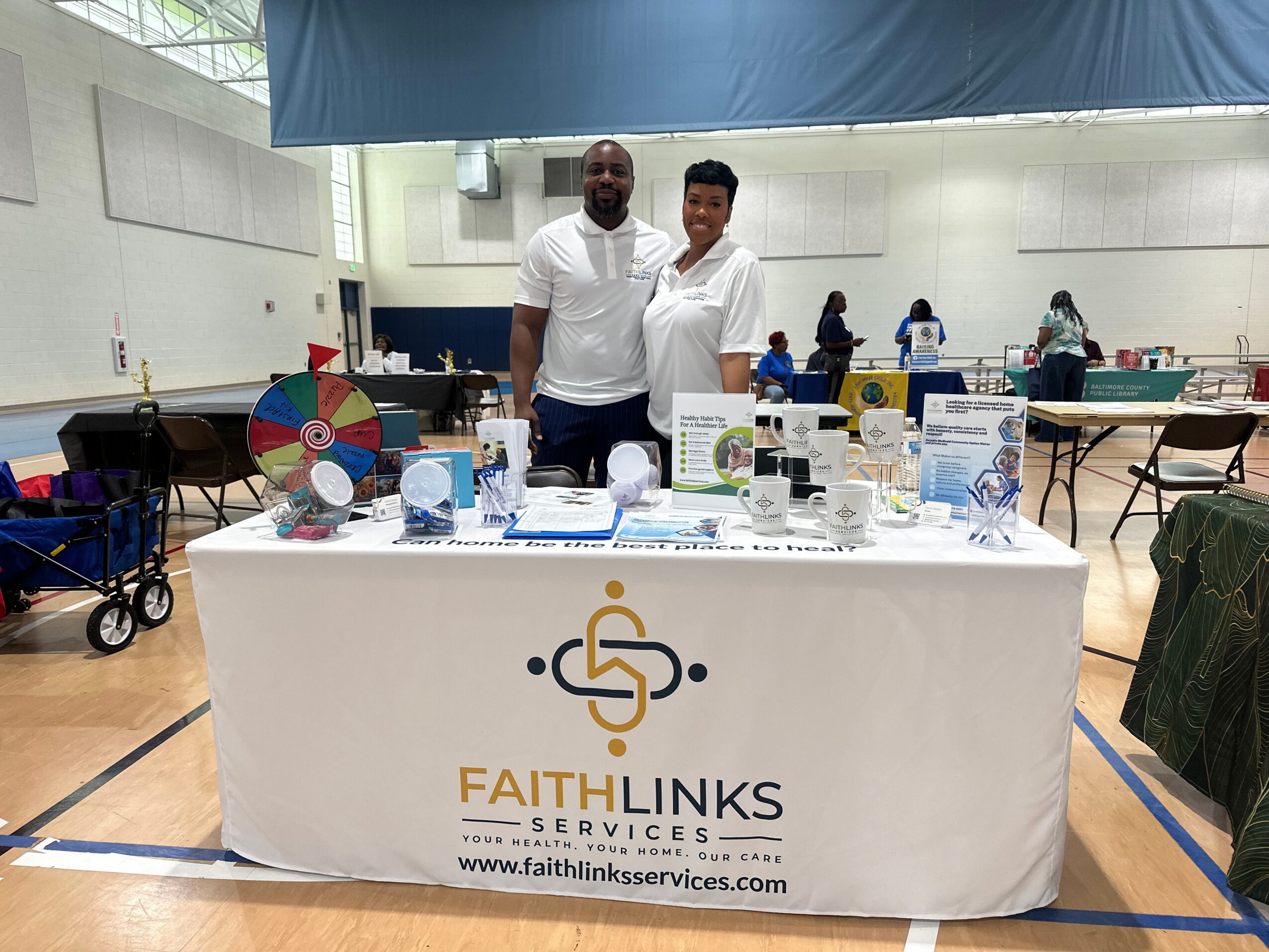 Two men stand behind a table with informational materials at an indoor event, with other tables and people in the background.