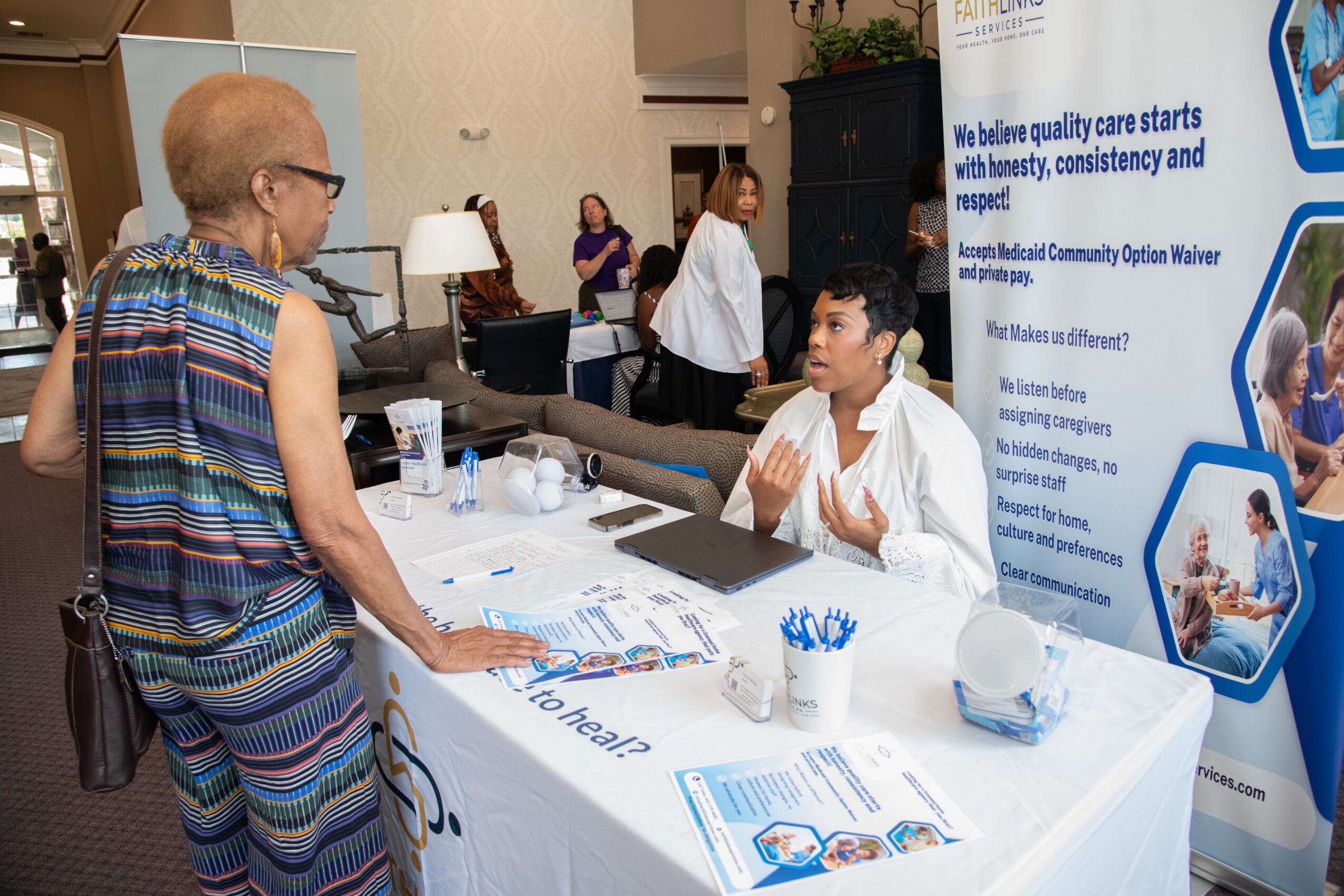 A woman in colorful striped clothing talks to a man seated at a table with informational materials, in front of a banner. Word count: 20