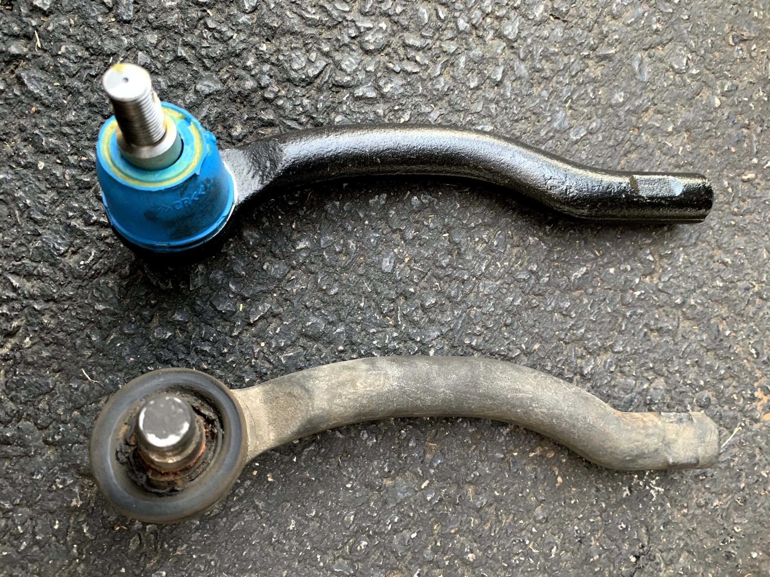 Two car steering tie rods, one black and one gray, placed on a textured surface.