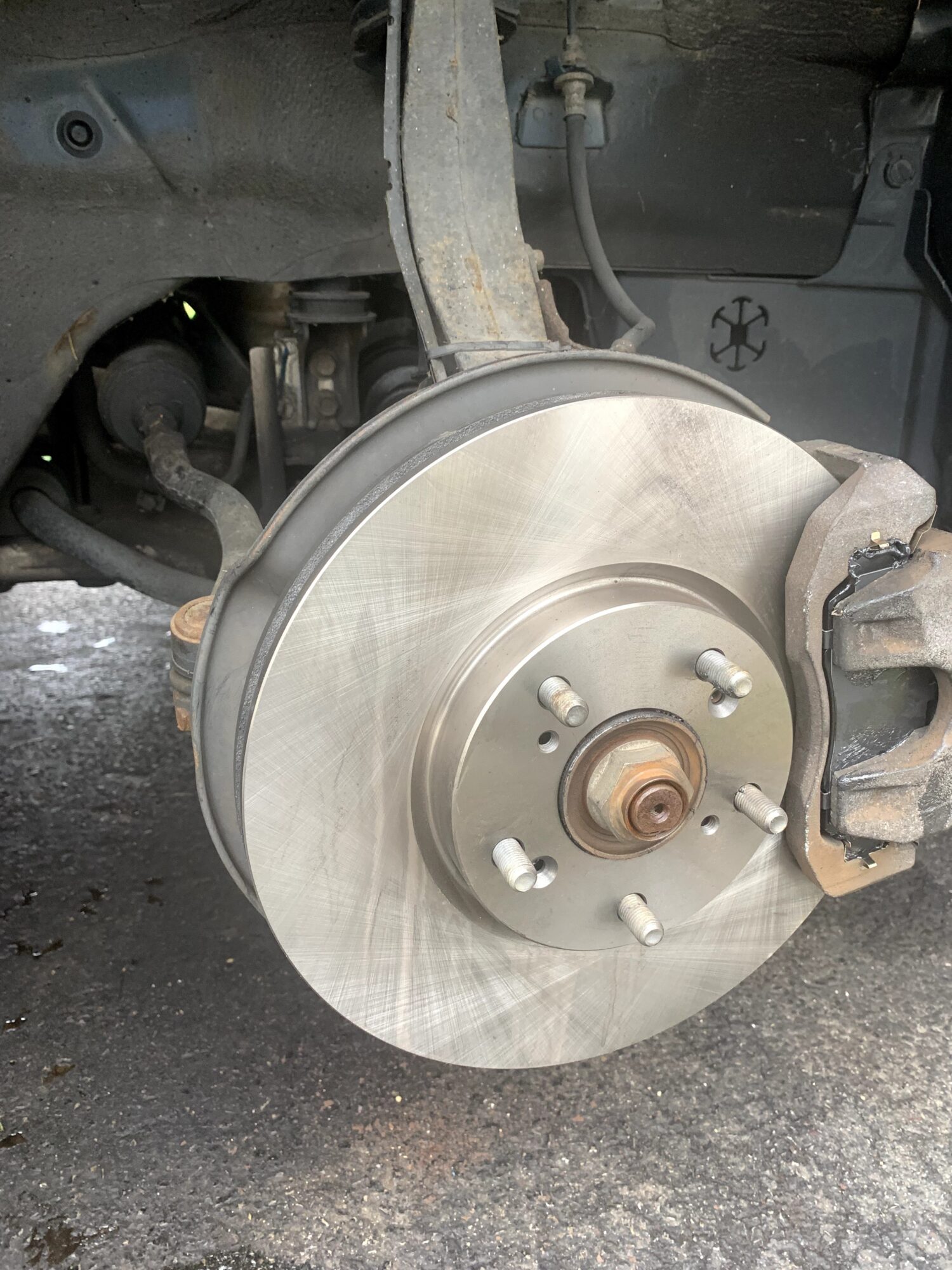 Car's brake disc and caliper assembly visible, with wheel removed, on a paved surface.