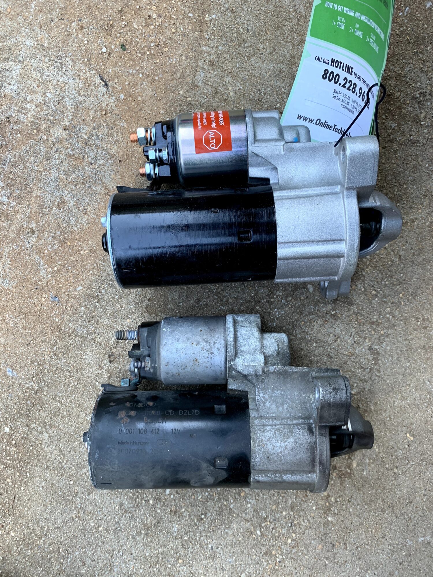 Two automotive starters placed on a concrete surface, one silver and black, the other gray and black.