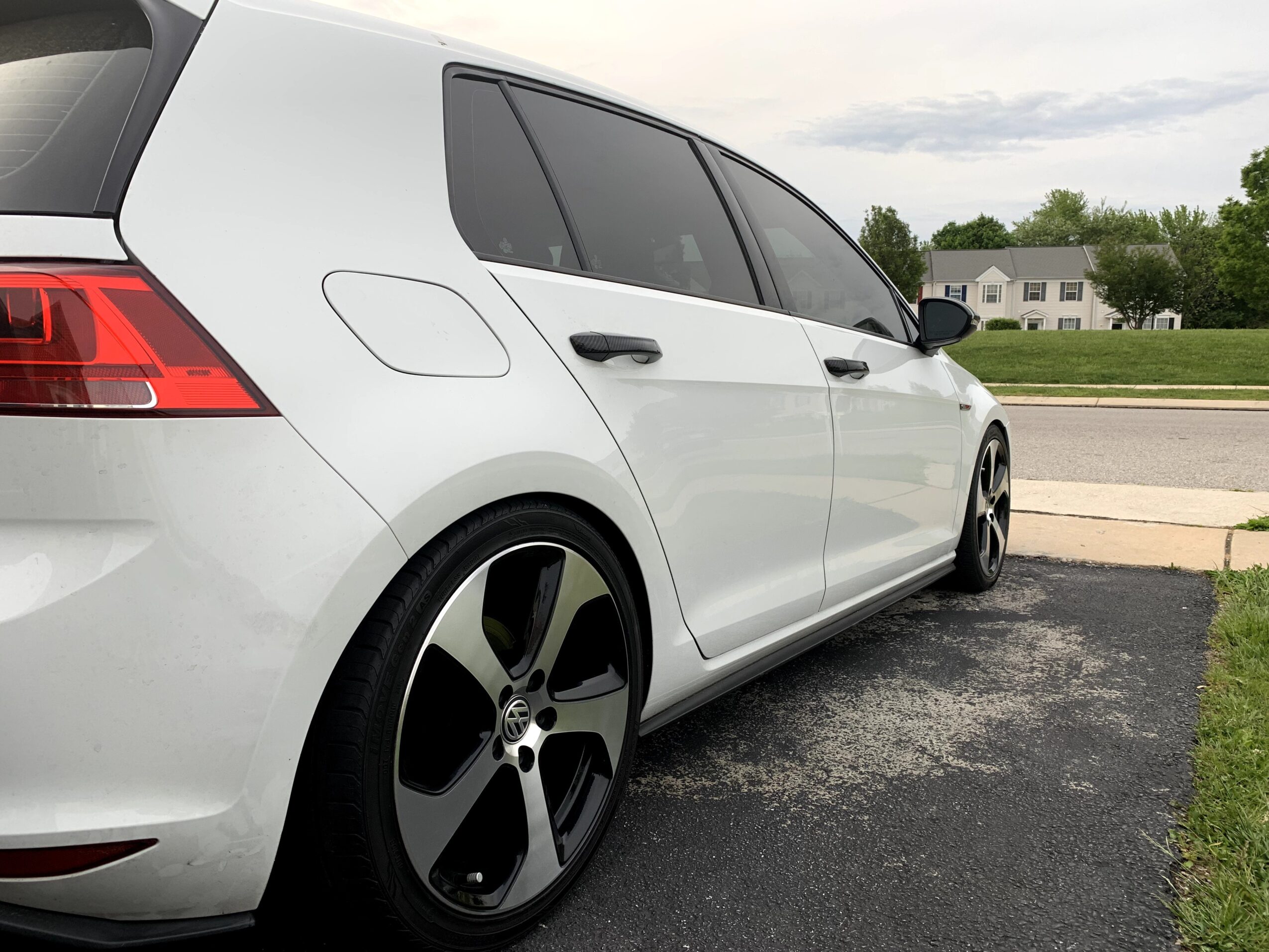 White hatchback car parked on driveway, with houses and trees in background.