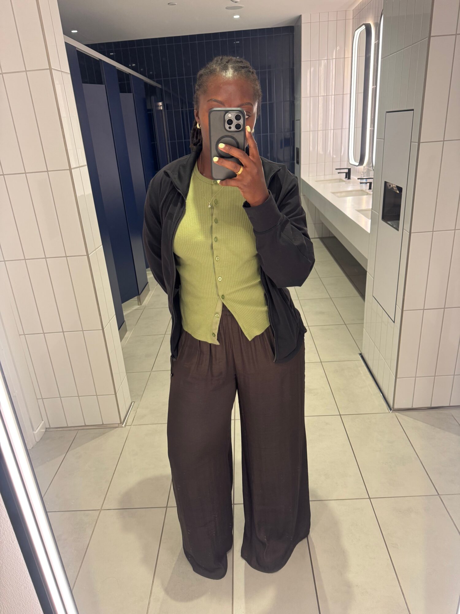 Person taking a mirror selfie in a bathroom, wearing a black jacket and green shirt, holding a phone with a triple-lens camera, standing on a tiled floor.