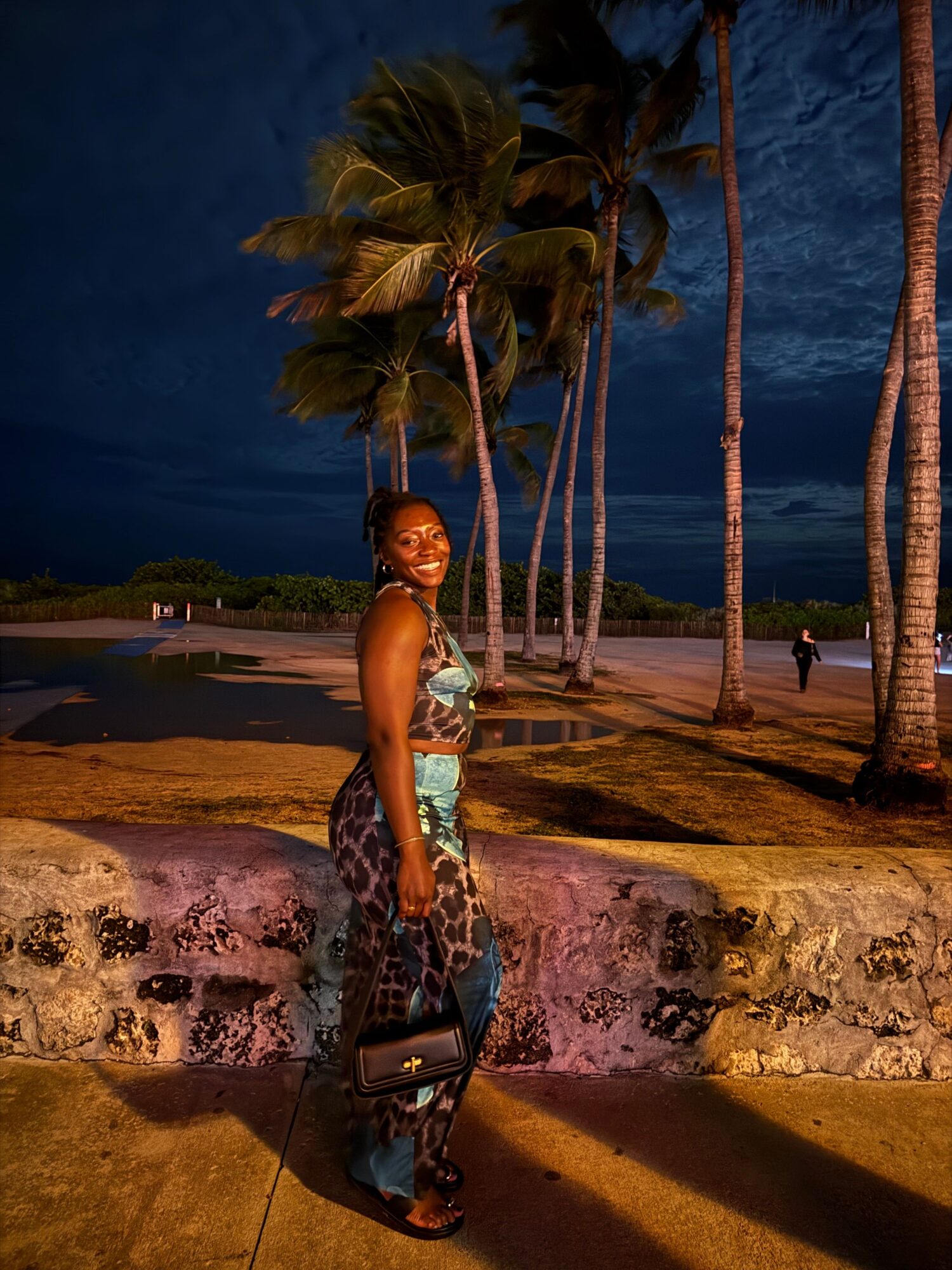 Person standing on a sandy beach with palm trees and water in the background, wearing a patterned outfit and sandals.