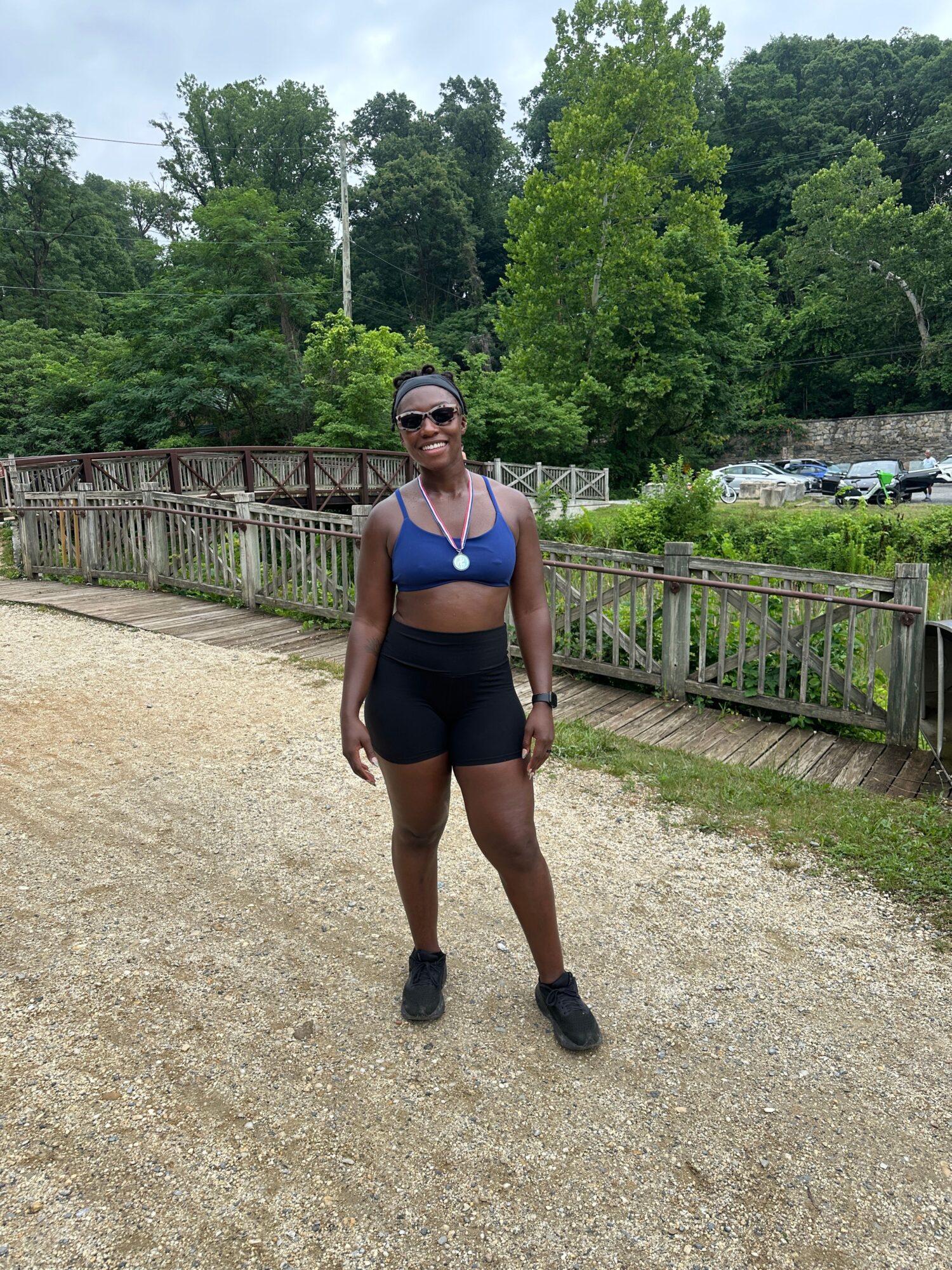 Woman standing outdoors on a gravel path, wearing sunglasses, a blue top, and black shorts, with trees and a wooden fence behind her.