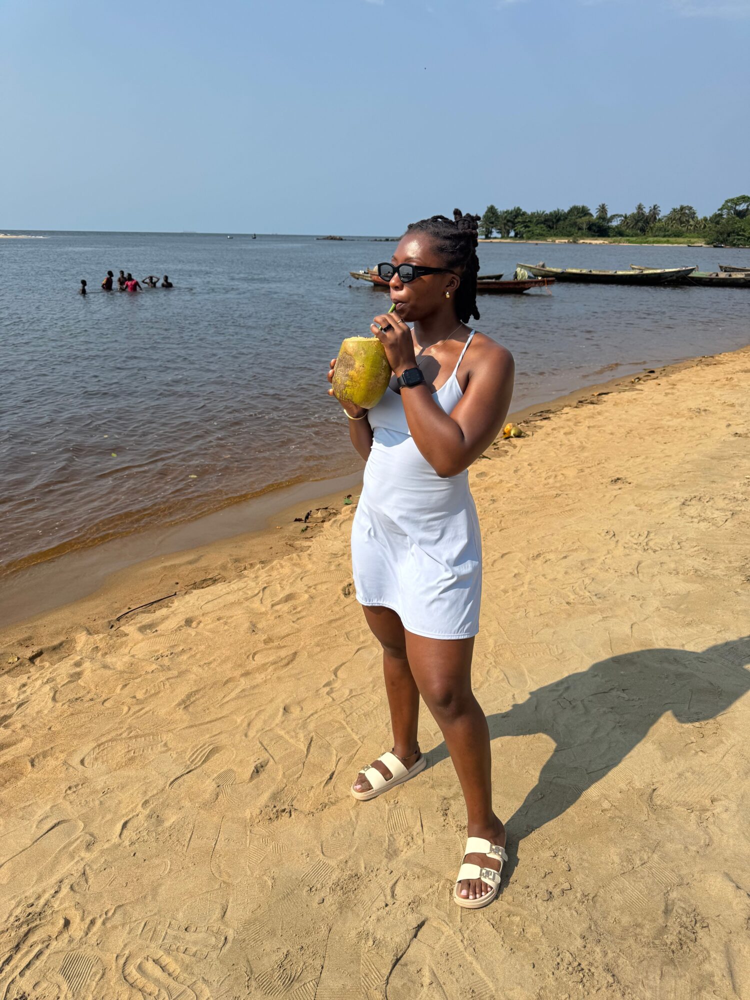 Person standing on sandy beach near water, holding a yellow drink, wearing a white dress and sandals, with a shadow cast on sand.