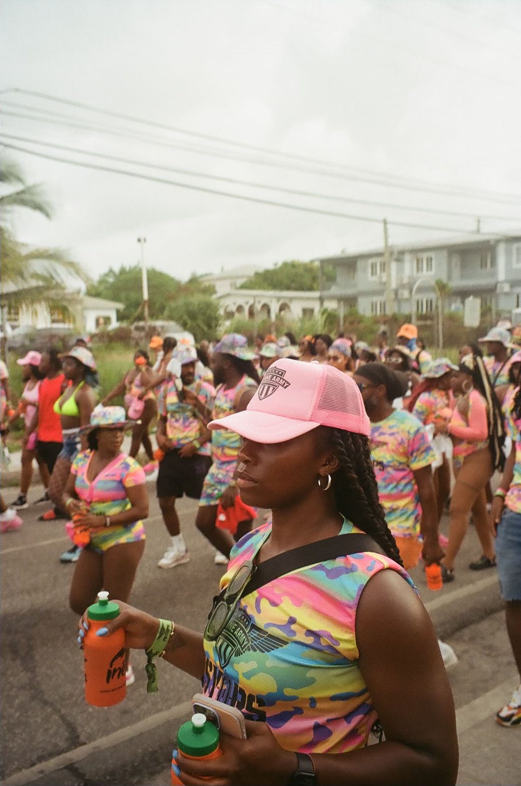 Crowd of people walking outdoors, some wearing colorful clothing and hats, with power lines and trees in the background.