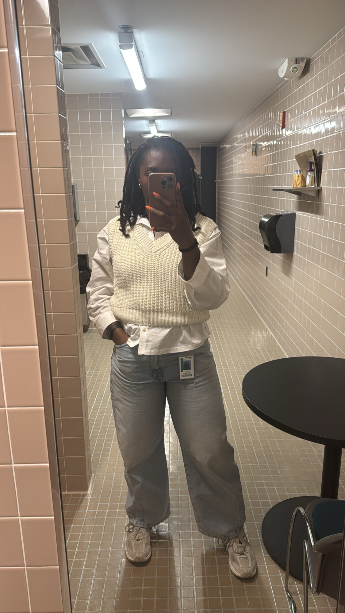 Person taking a mirror selfie in a bathroom, wearing a white sweater, jeans, and sneakers.