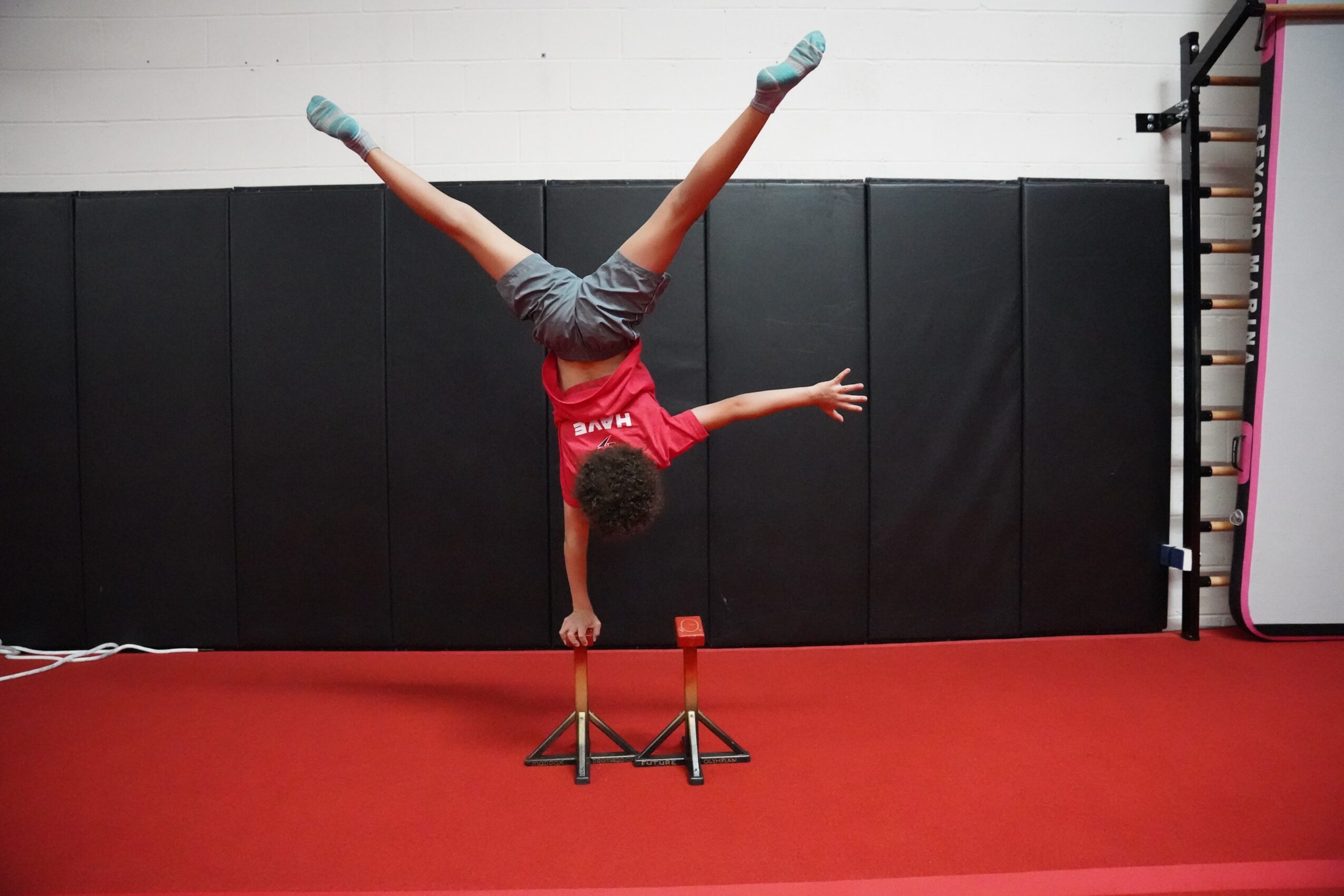 Person performing a handstand with legs spread, balancing on hands, in a gym with black padded walls and red floor.