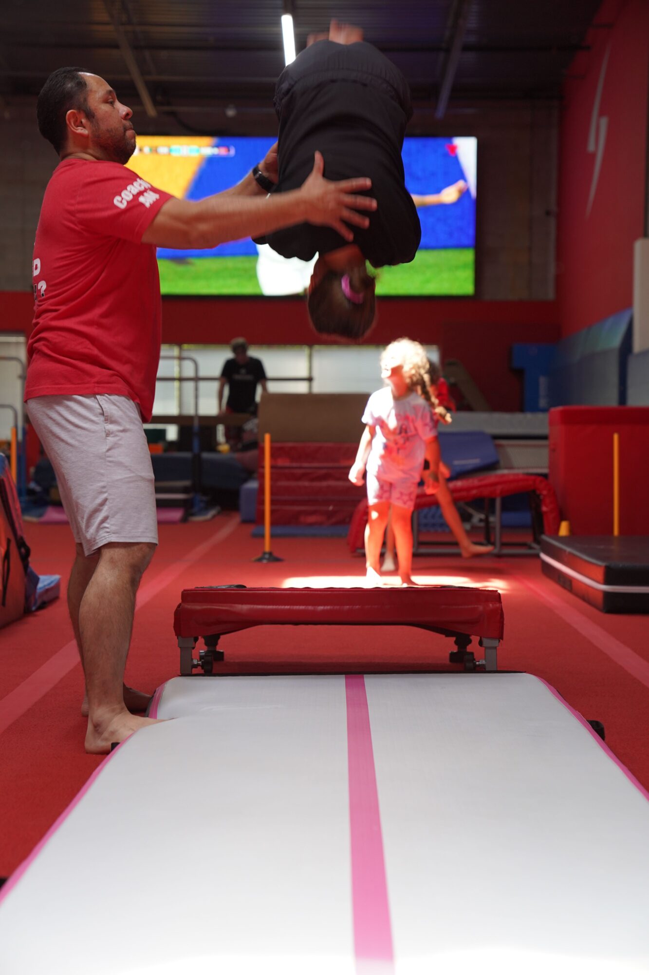 Two men in a gym, one giving a massage to the other, with gym equipment and a TV screen in the background.