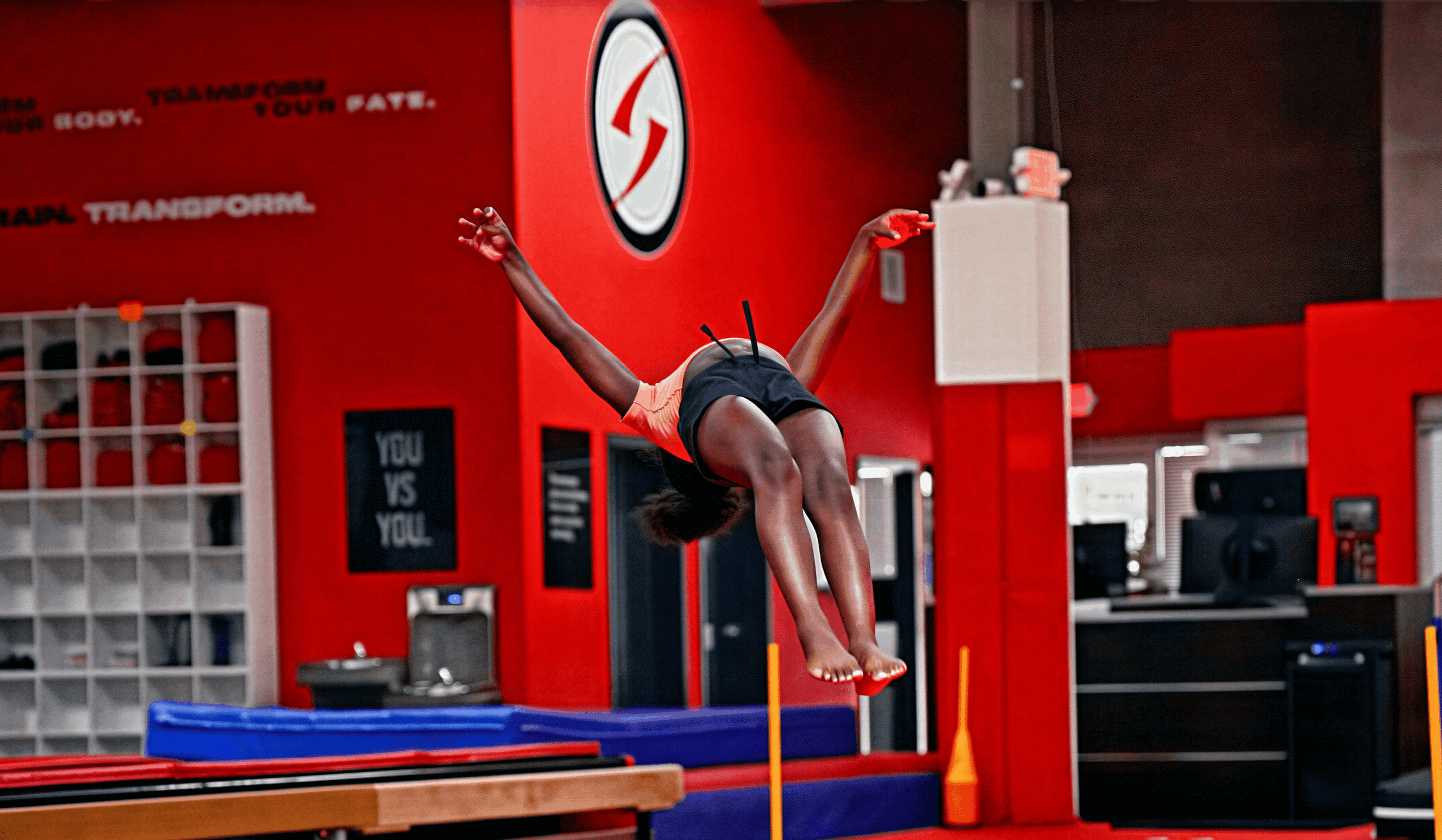 A gymnast in mid-air during a vault, with a red and black background and gym equipment visible.