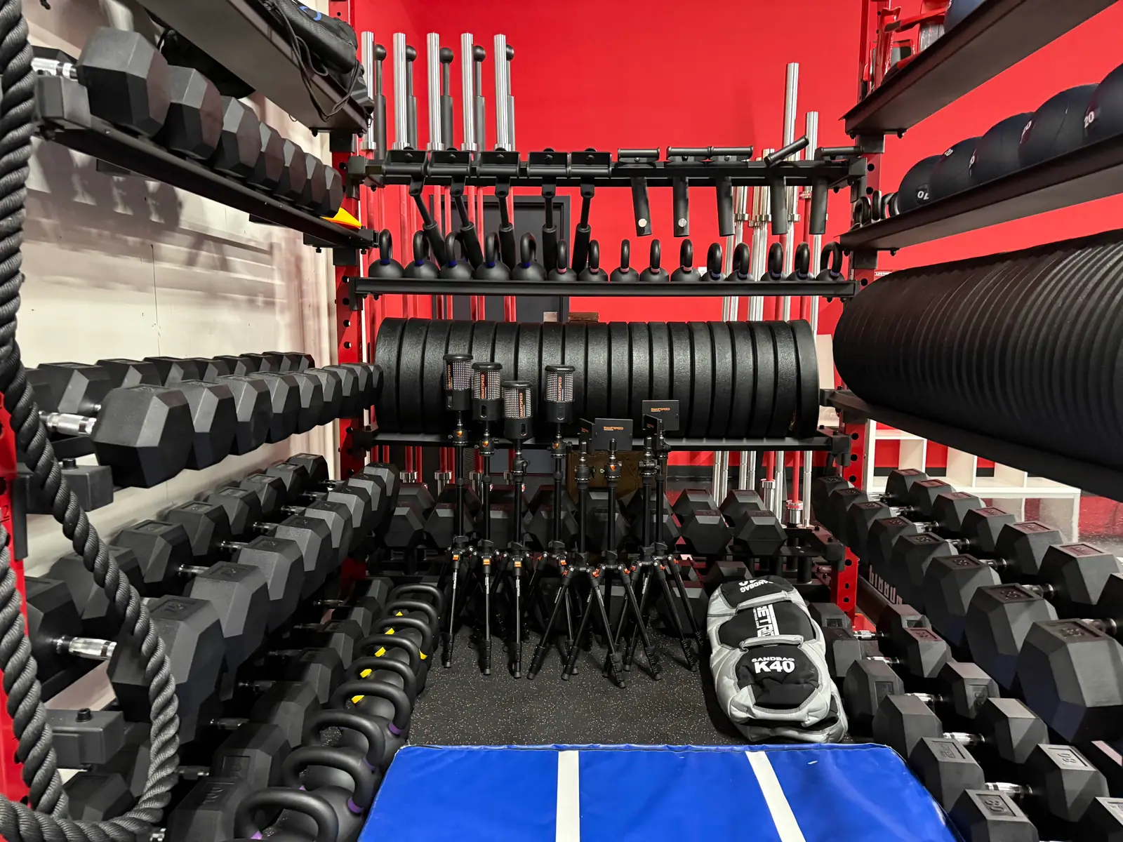 Gym equipment storage with kettlebells, dumbbells, and resistance bands organized on shelves and racks, with a blue mat on the floor.