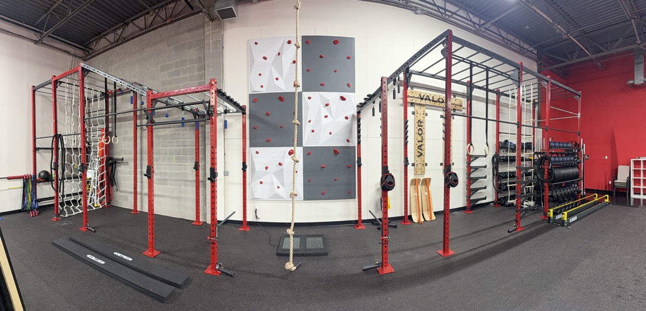 Indoor gym with red and black climbing structures, hanging ropes, and workout equipment against a white wall.