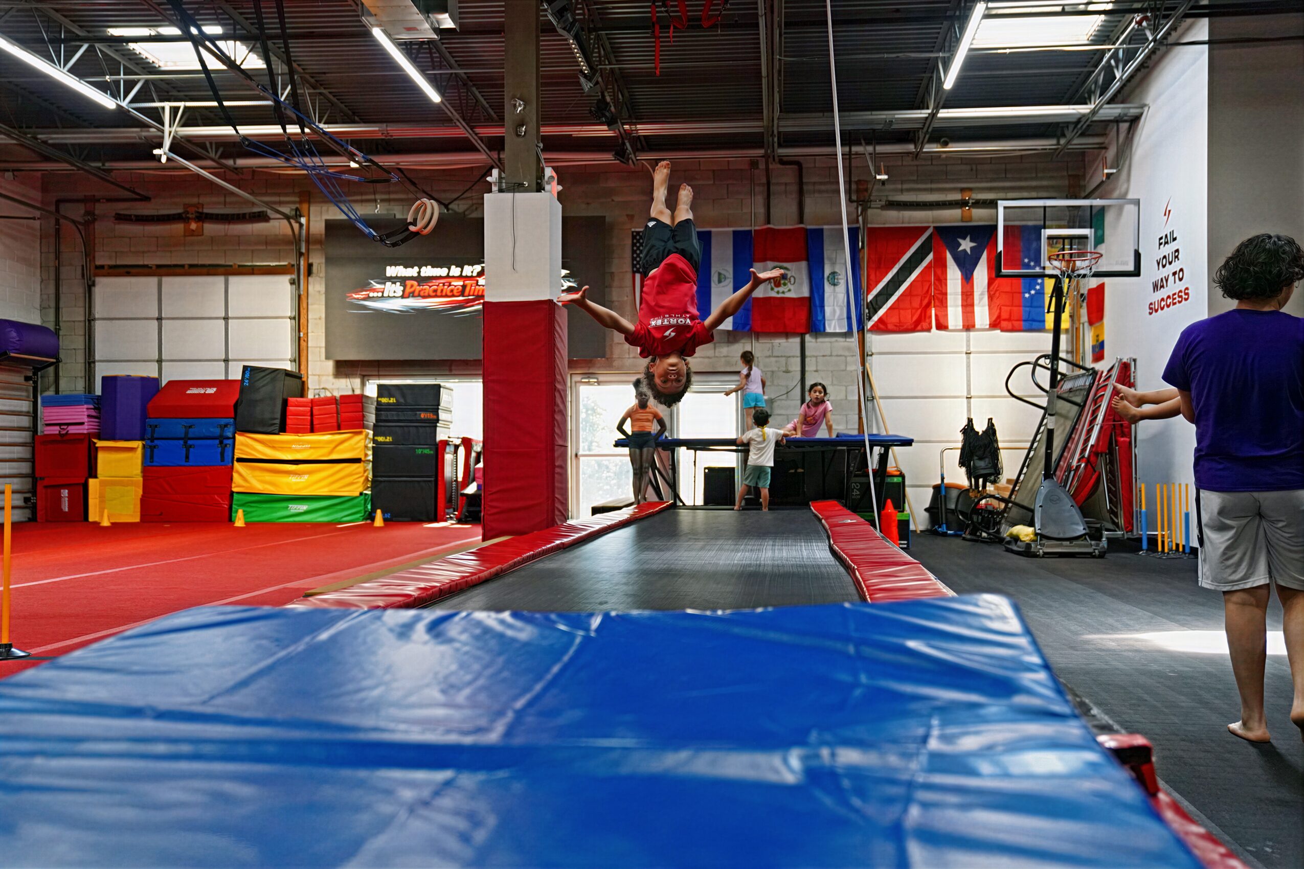 Person performing a flip on a trampoline in an indoor gym with equipment and people watching.