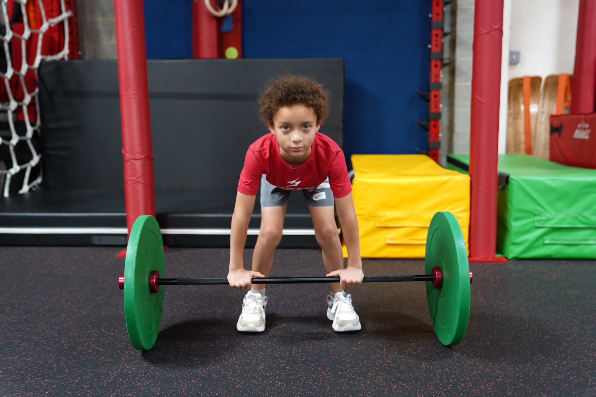 Child with curly hair lifting a barbell with green weights in a gym, colorful mats and equipment in background.