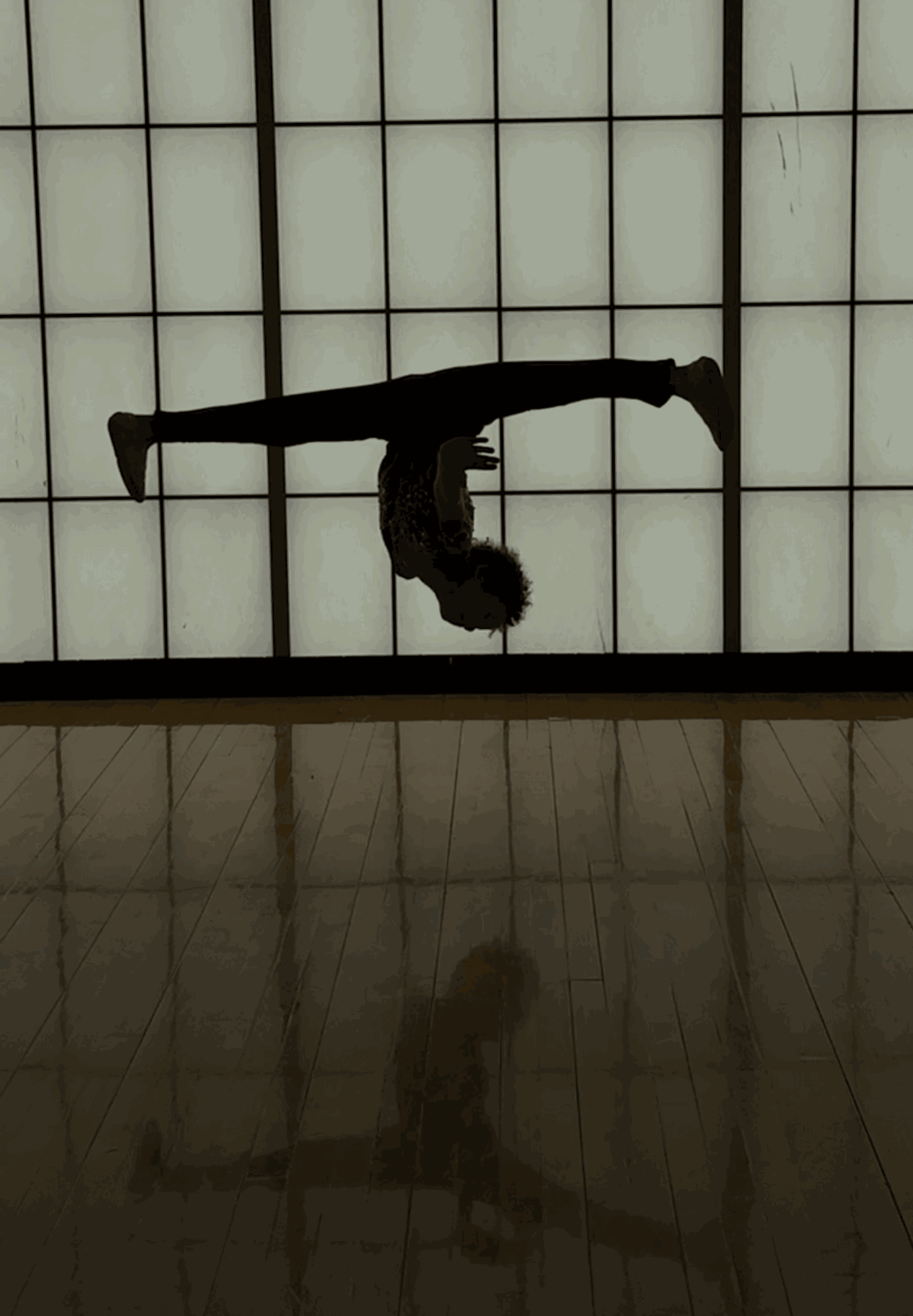 Person performing a backbend with arms and legs extended, silhouette against a backlit grid wall, reflection on shiny floor.