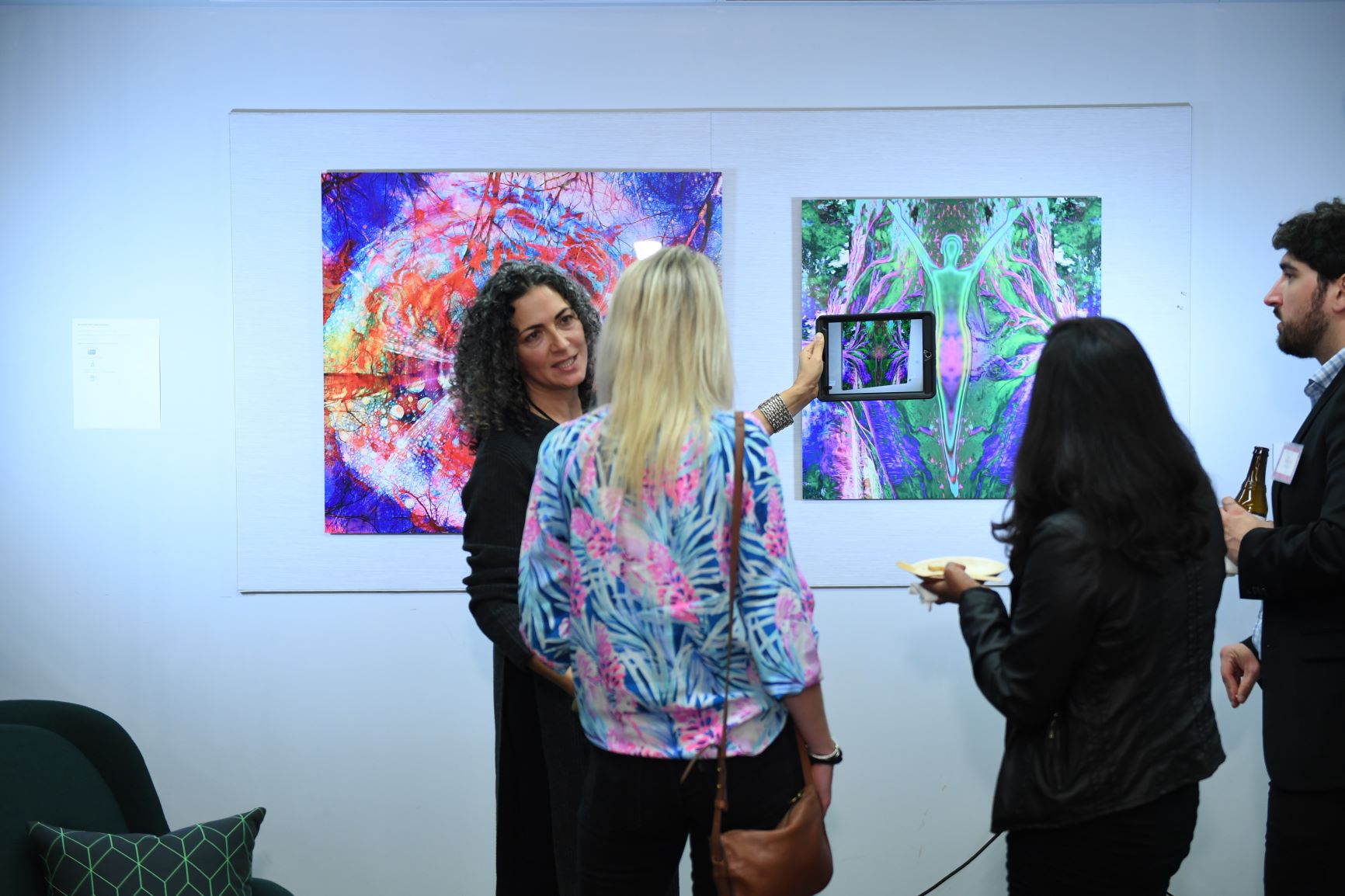 Four people in an art gallery looking at colorful abstract paintings, one person taking a photo with a tablet.