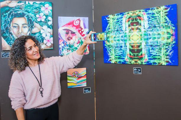 Woman with curly hair takes a photo of colorful digital art on a wall, smiling. Art features a face with vibrant patterns.