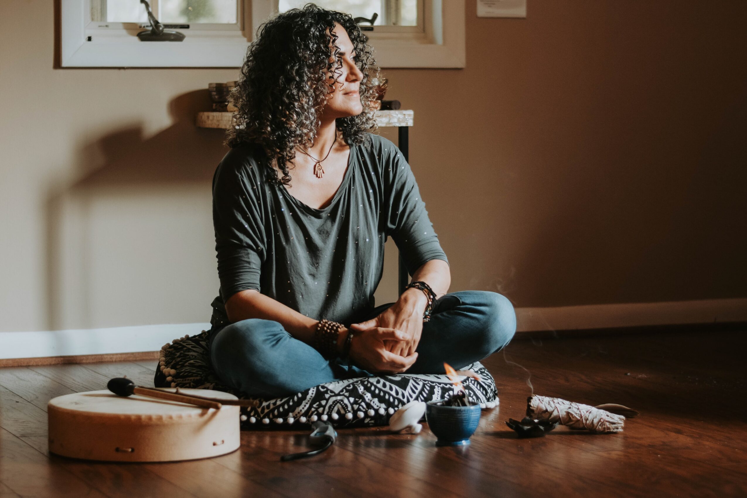 Woman with curly hair sitting cross-legged on a cushion, looking to the side, with sunlight coming through a window behind her.