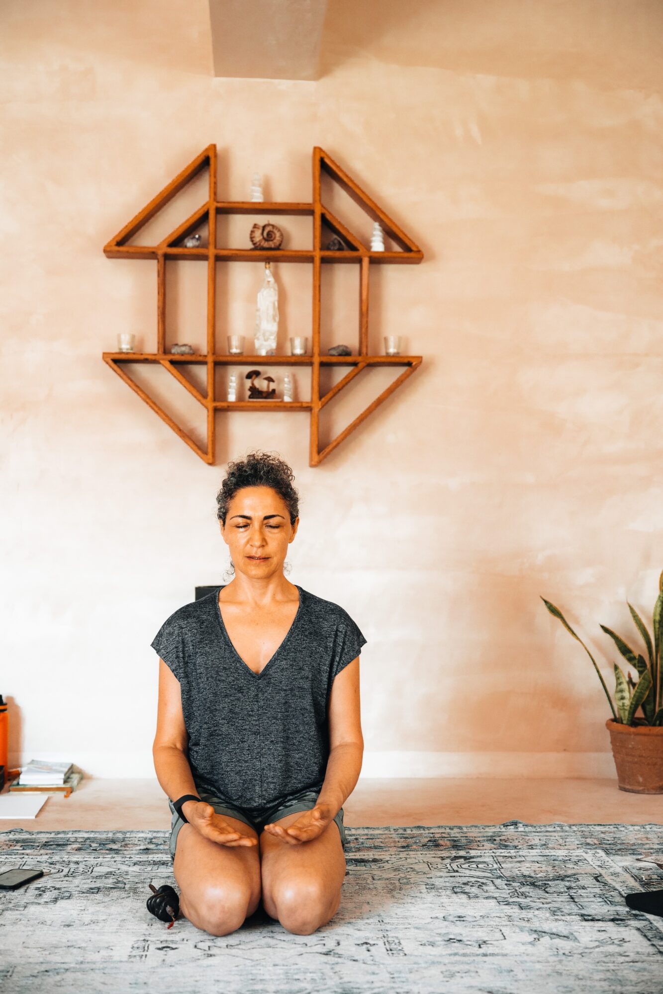 Woman sitting cross-legged on a rug with a house-shaped wooden shelf and potted plant behind her.