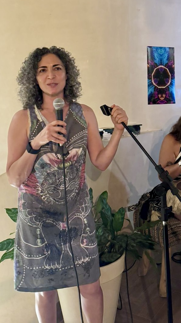 Woman with curly hair holding a microphone and a stand, standing indoors near a colorful artwork and plants.