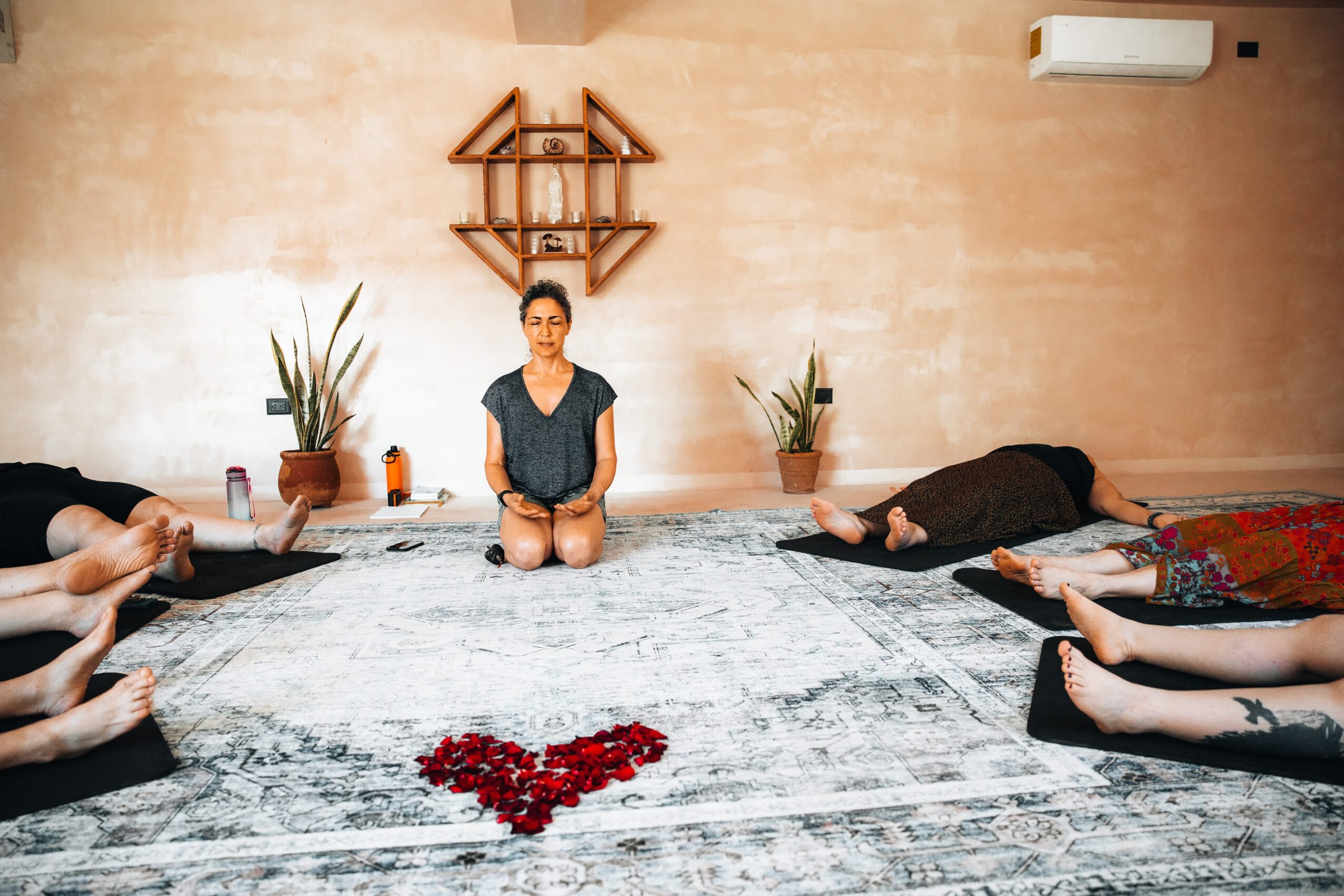 People participating in a yoga or meditation session on mats in a room with a beige wall, plants, and a heart-shaped arrangement of red objects on the rug.