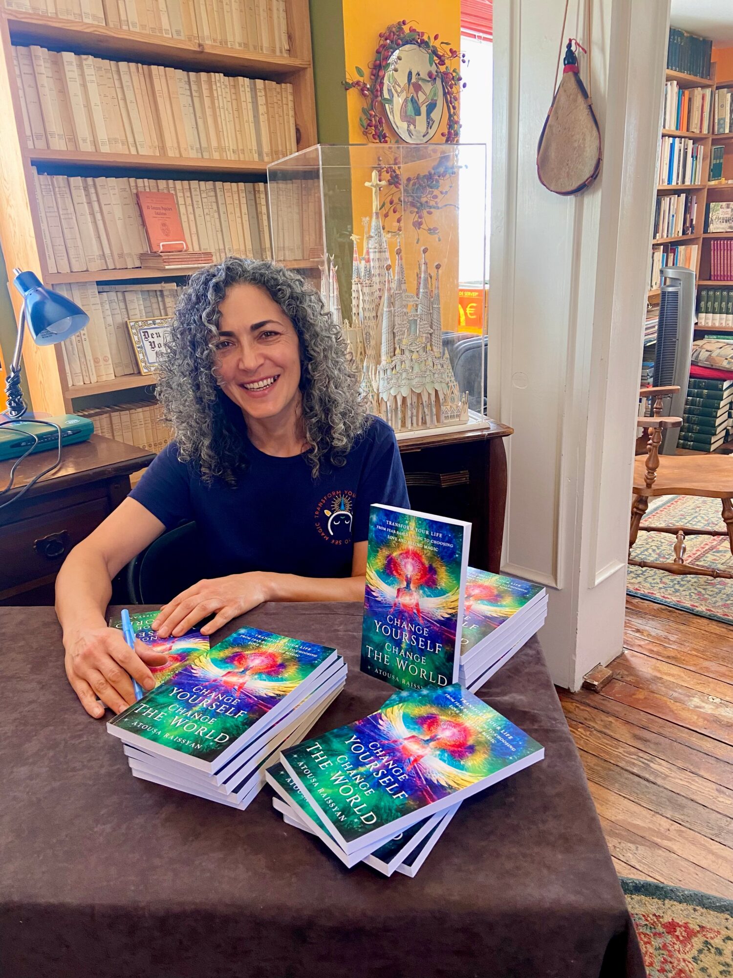 Woman with curly hair smiling at a table with colorful books in a cozy library setting.