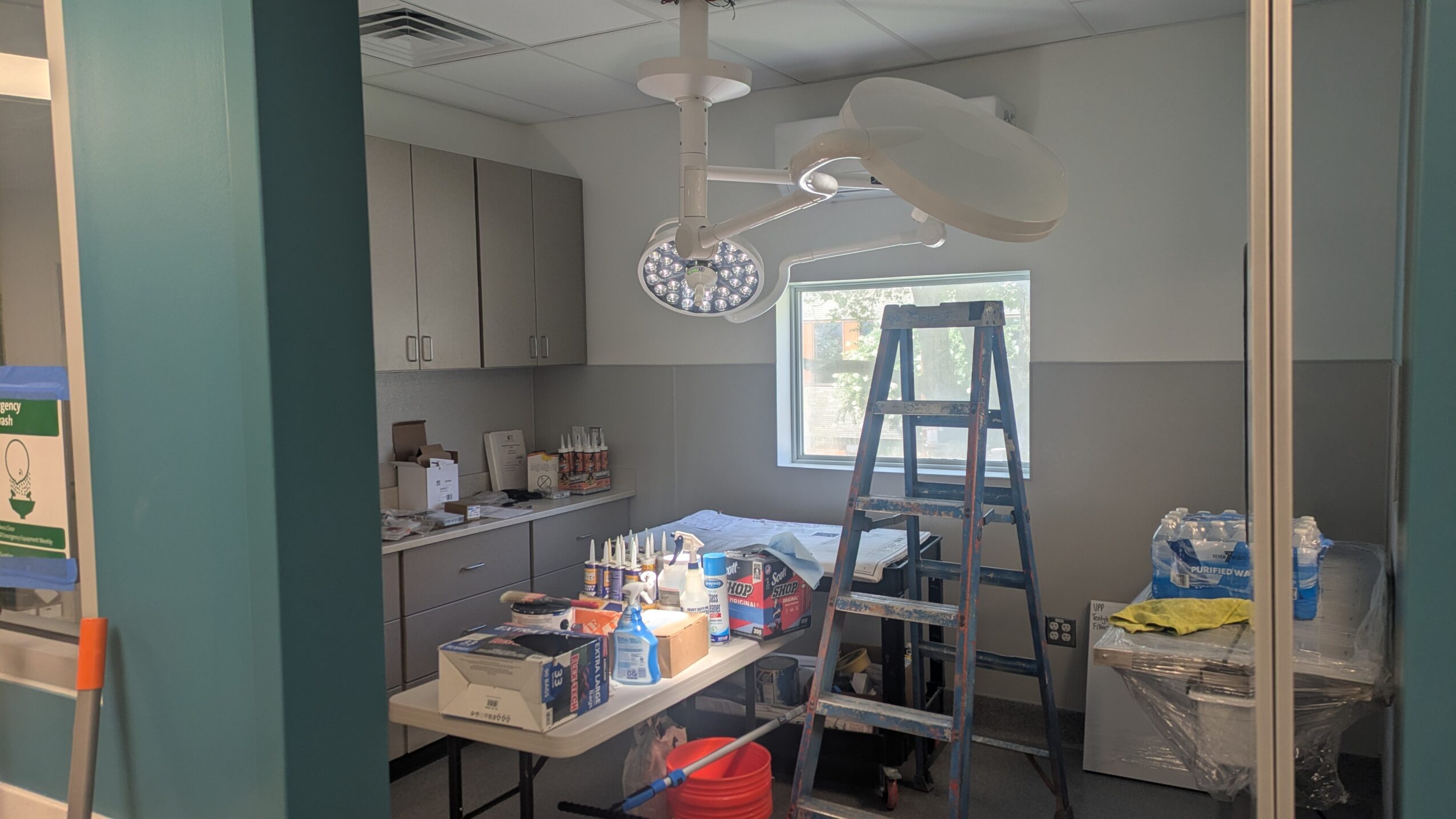 Medical room with cabinets, a table with supplies, a ladder, and a window, viewed through glass doors.